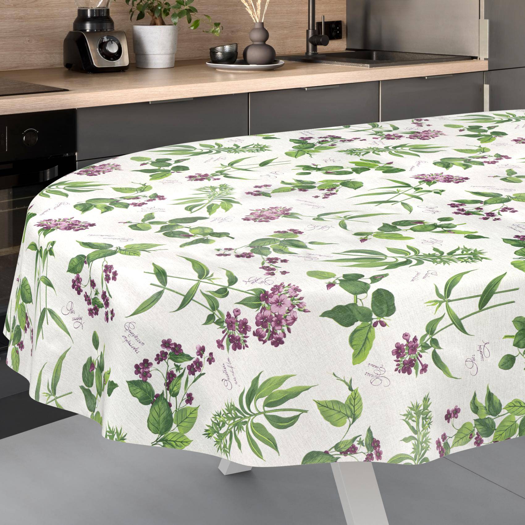 Ein runder Tisch mit einer weißen Tischdecke mit Blumenmuster, auf der grüne Blätter und violette Blüten in einer modernen Küche zu sehen sind.
