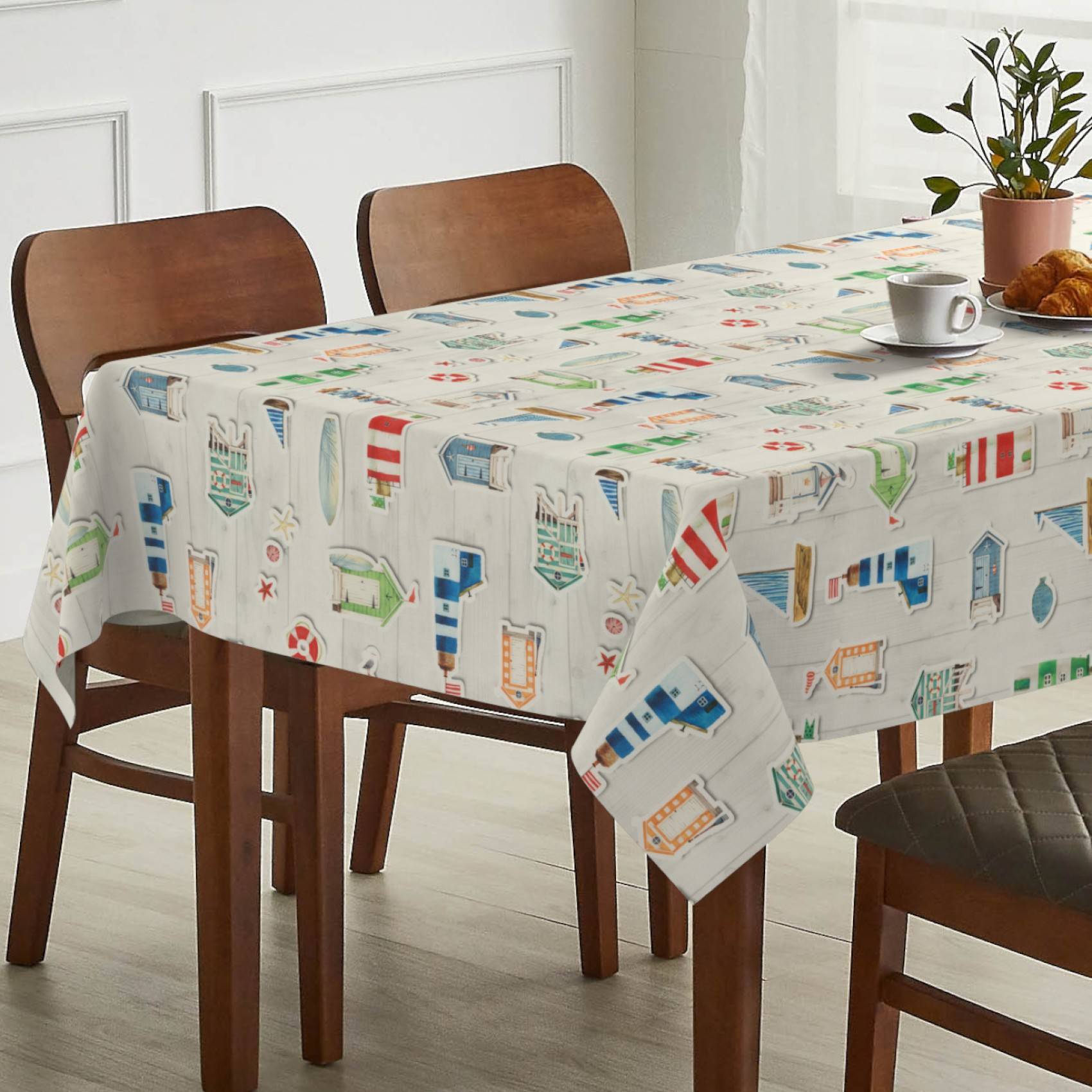 Esstisch mit einer Tischdecke mit Leuchtturmuster, eingedeckt mit einer Tasse, einem Croissant und einer getopften Pflanze neben Holzstühlen in einem hellen Raum.