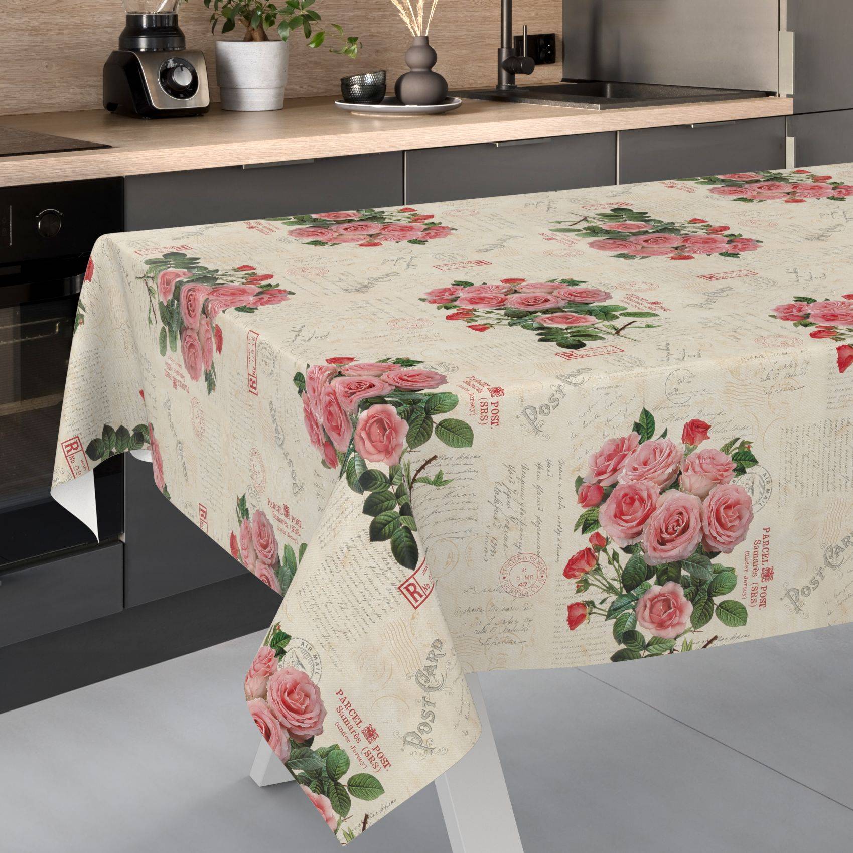Küchentisch bedeckt mit einer cremefarbenen Blümchentischdecke, bedruckt mit rosa Rosen und Vintage-Postkartentexten.