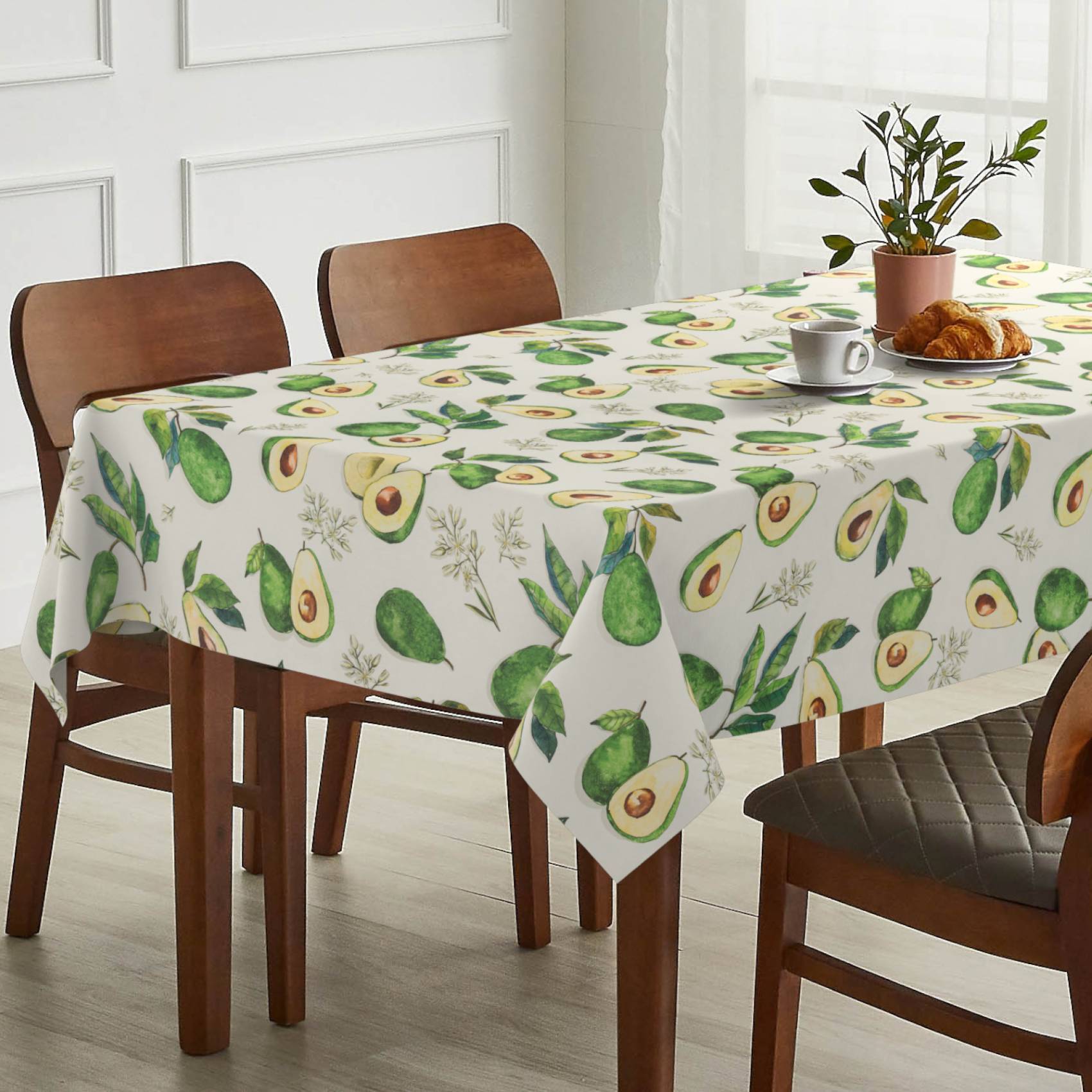 Esstisch mit avocadomusterter Tischdecke, eingedeckt mit Kaffee, Gebäck und einer Topfpflanze in einem hellen Zimmer.