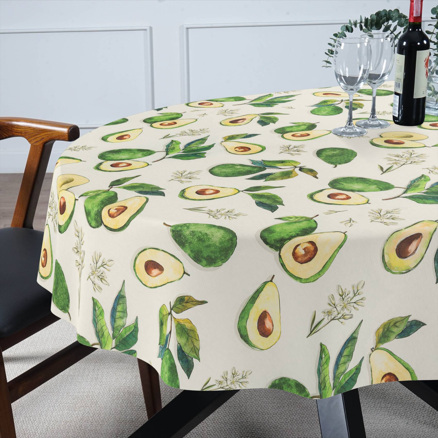 Esstisch mit einer Tischdecke mit Avocado-Muster, eingedeckt mit Weingläsern und einer Flasche, in einem hellen Raum.