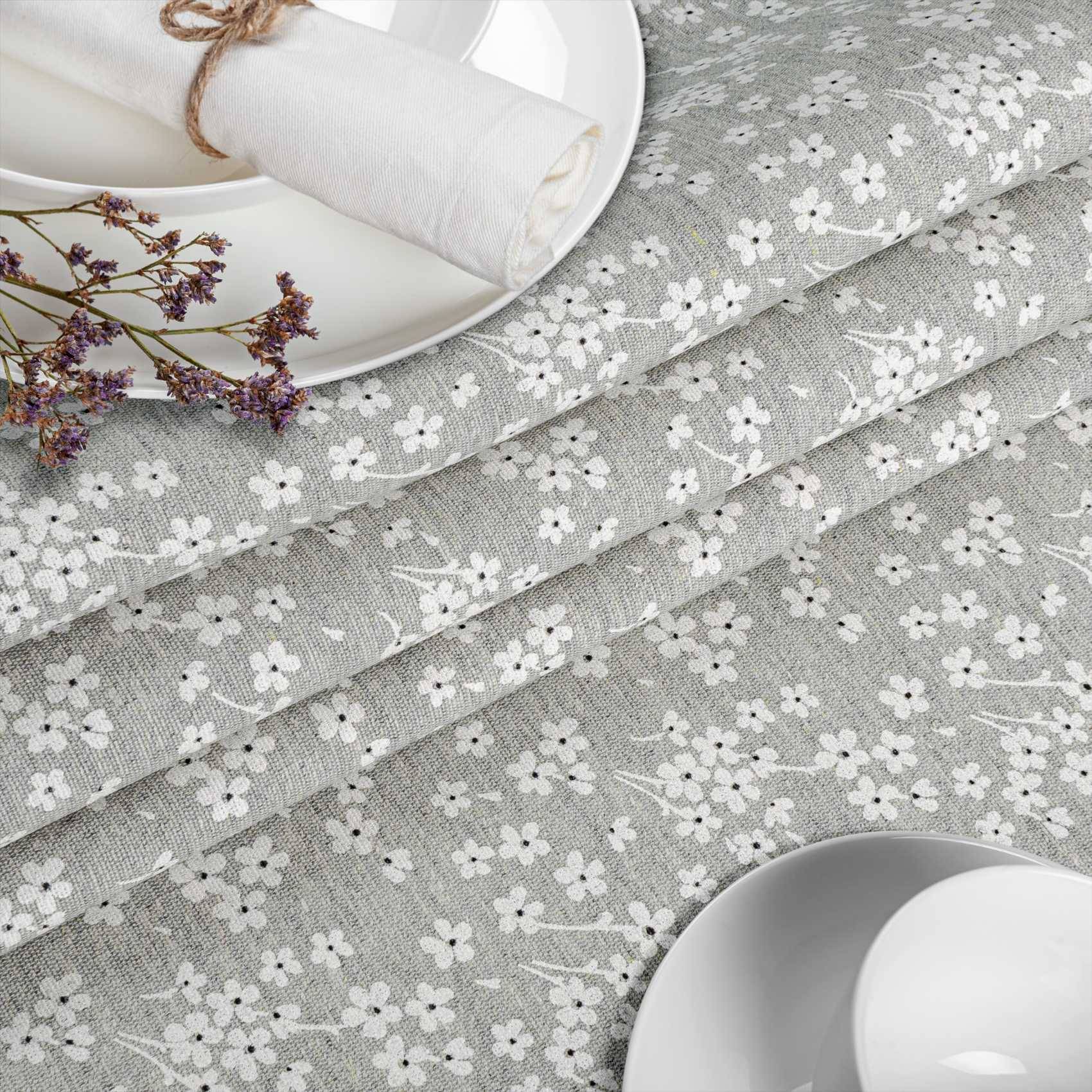 Graues Blumenmuster-Textil mit weißen Blüten, in gefalteten Schichten neben weißem Geschirr und einer zusammengerollten Serviette angeordnet, was auf Tischwäsche oder Textilmaterial hindeutet.