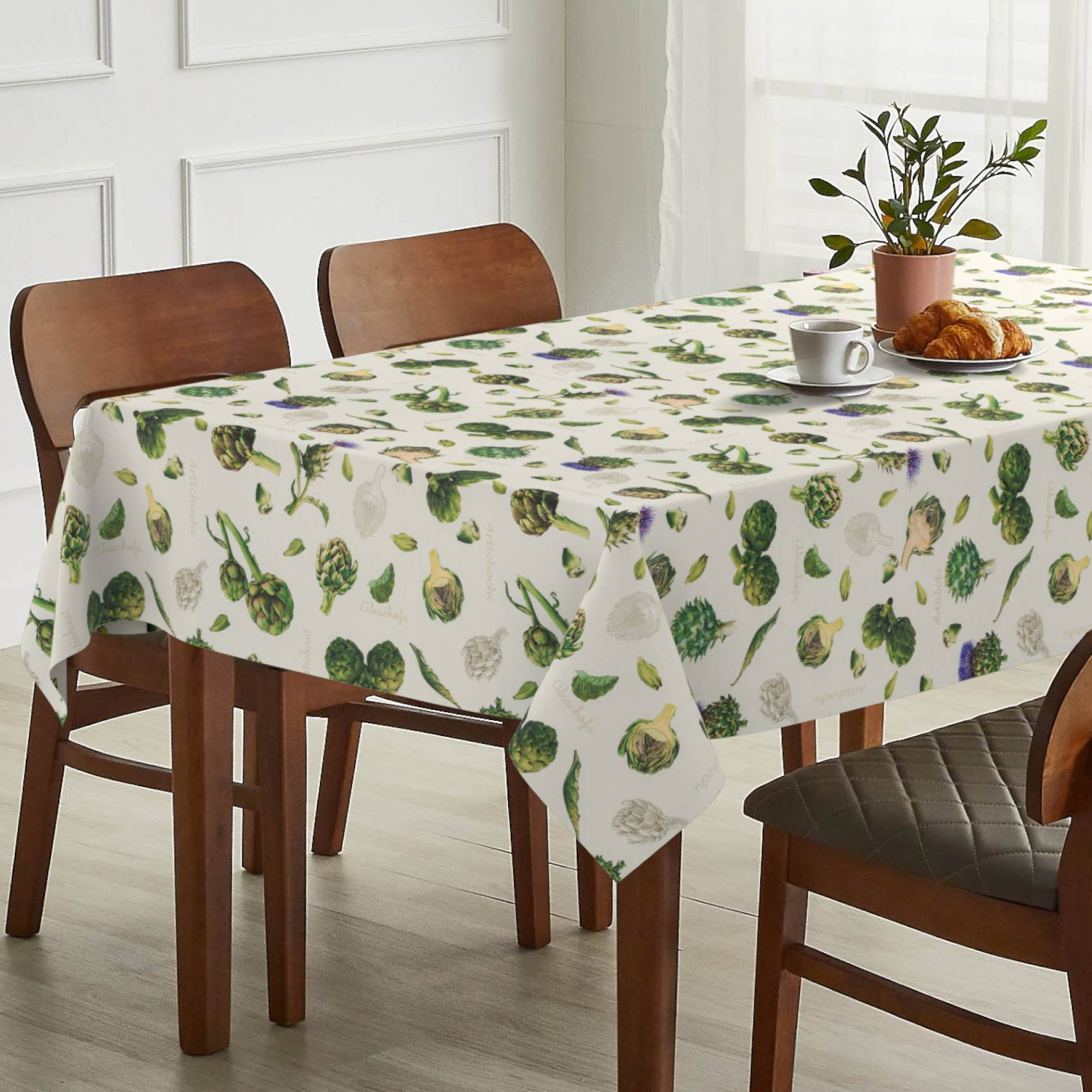 Esstisch mit einer weißen Tischdecke mit Blattmuster, eingedeckt mit einer Tasse, Croissants und einer Topfpflanze in einem hellen Raum.
