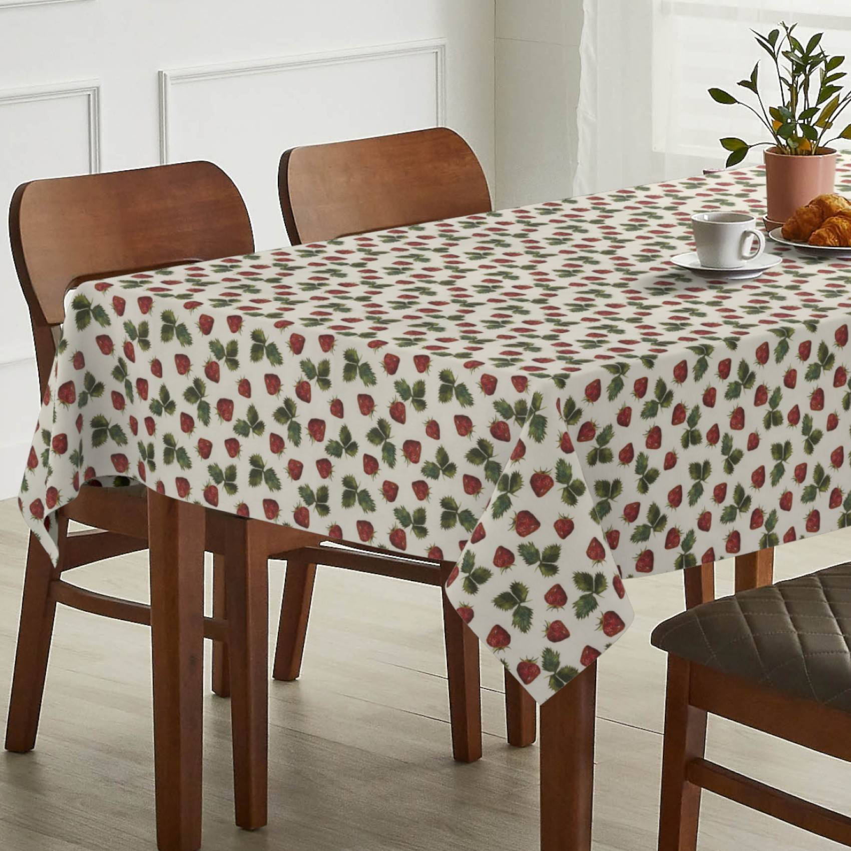 Esstisch mit erdbeergemusterter Tischdecke, eingedeckt mit einer Tasse, einem Croissant und einer Topfpflanze in einem hellen Raum.