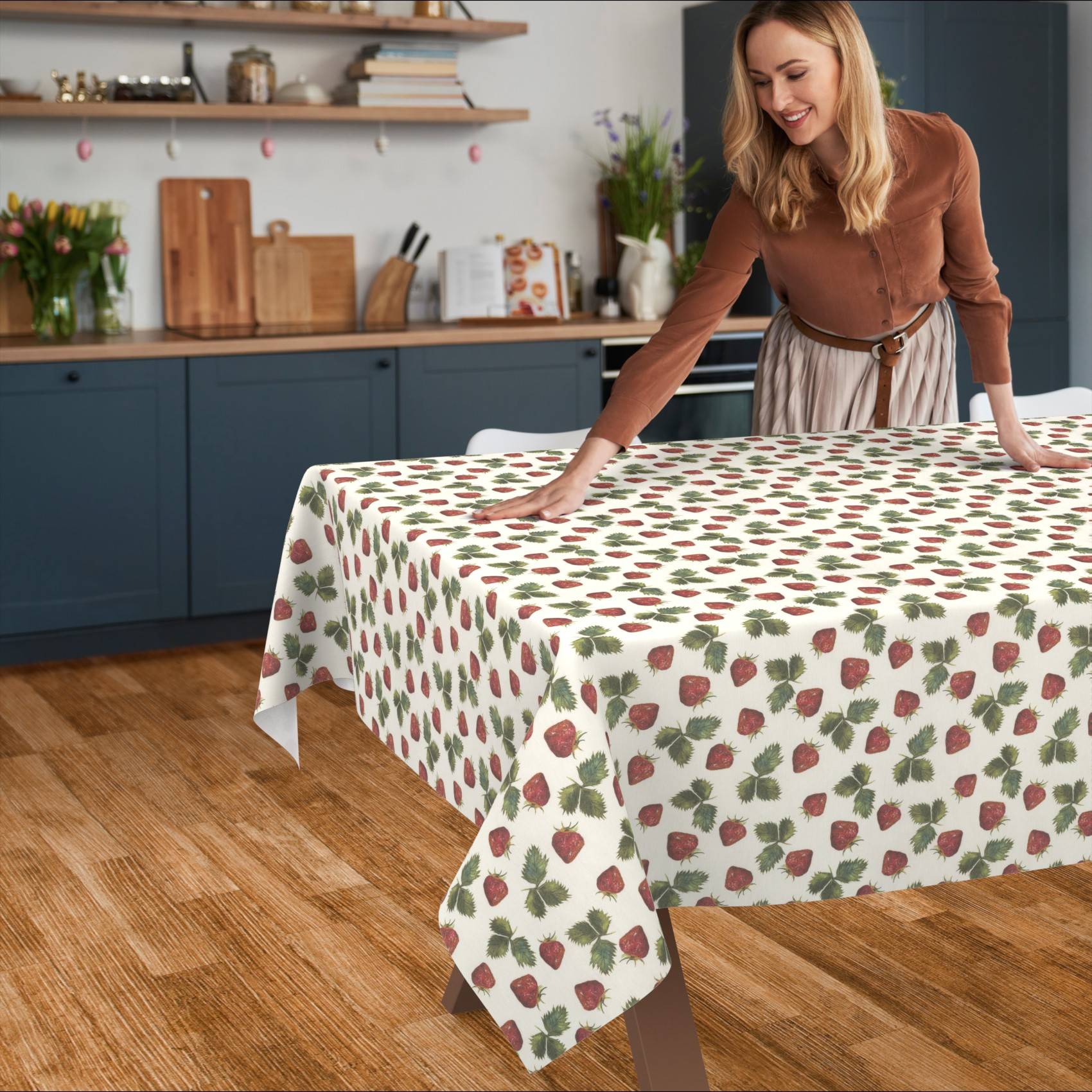 Eine Frau glättet eine Tischdecke mit Erdbeermuster über einem Esstisch in einer Küche.
