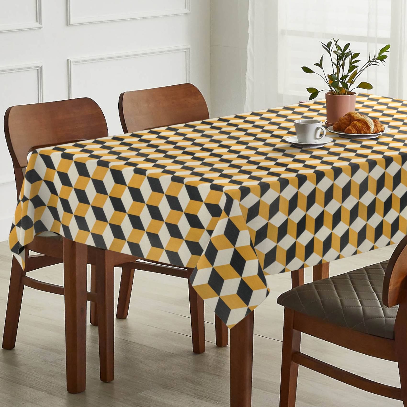 Esstisch mit geometrischem gelb-schwarz-weißem Tischtuch, eingedeckt mit einer Tasse, einem Croissant und einer Topfpflanze neben Holzstühlen.