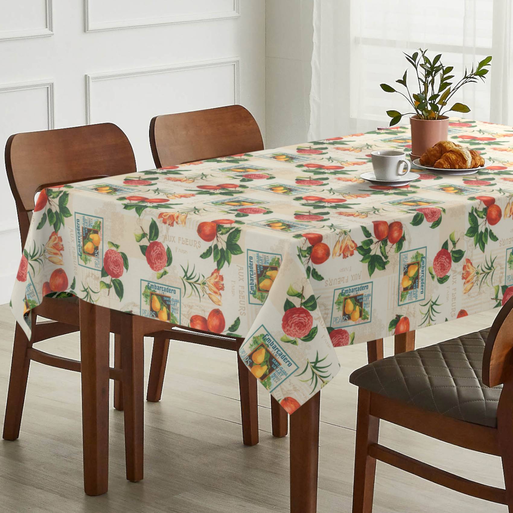 Esstisch mit Obstmuster-Tischdecke, eingedeckt mit einer Teetasse, einem Croissant und einer Topfpflanze neben Holzstühlen.