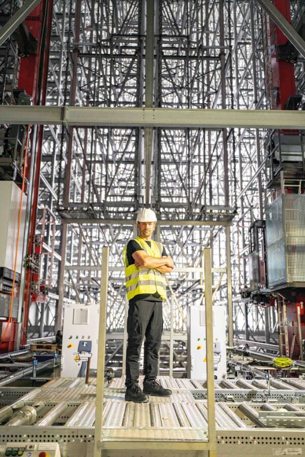 Arbeiter mit Schutzhelm und Warnweste steht auf einer Plattform innerhalb eines großen Stahlgerüsts oder einer Baustelle.