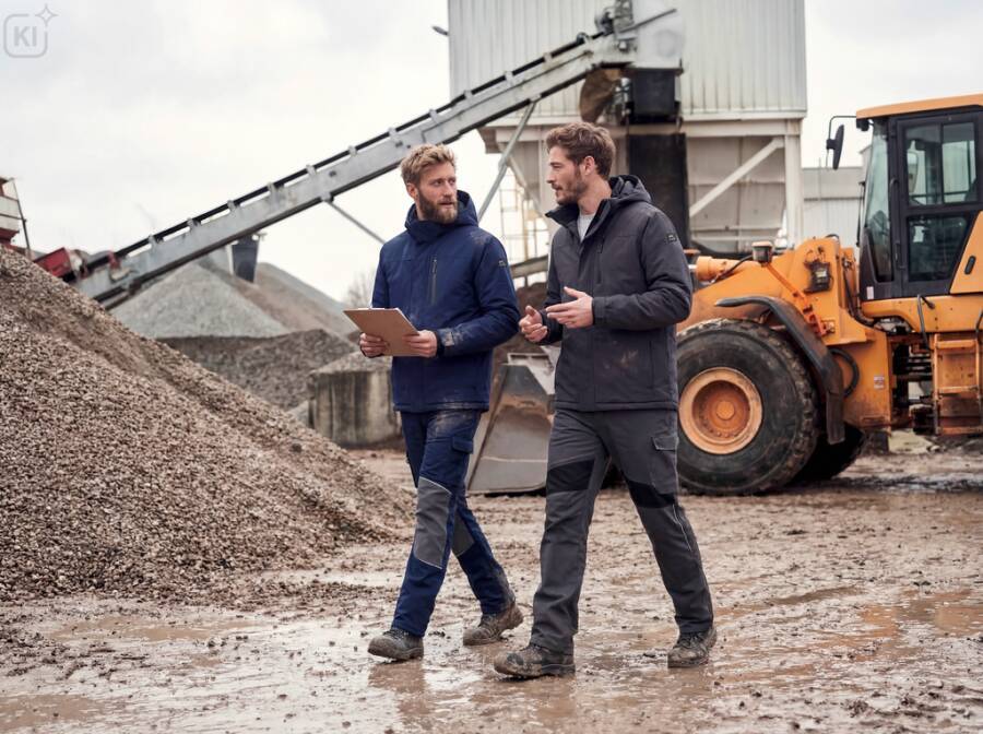 Zwei Männer in Outdoor-Arbeitskleidung besprechen eine Zwischenablage in einem Kieswerk, mit einem Bagger und einem Förderband im Hintergrund.