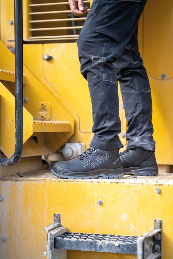 Ein Arbeiter steht auf gelbem Schwellastmaschinen, trägt schwarze Atlas-Sicherheitsstiefel und dunkle Hosen, gezeigt von der Taille abwärts.