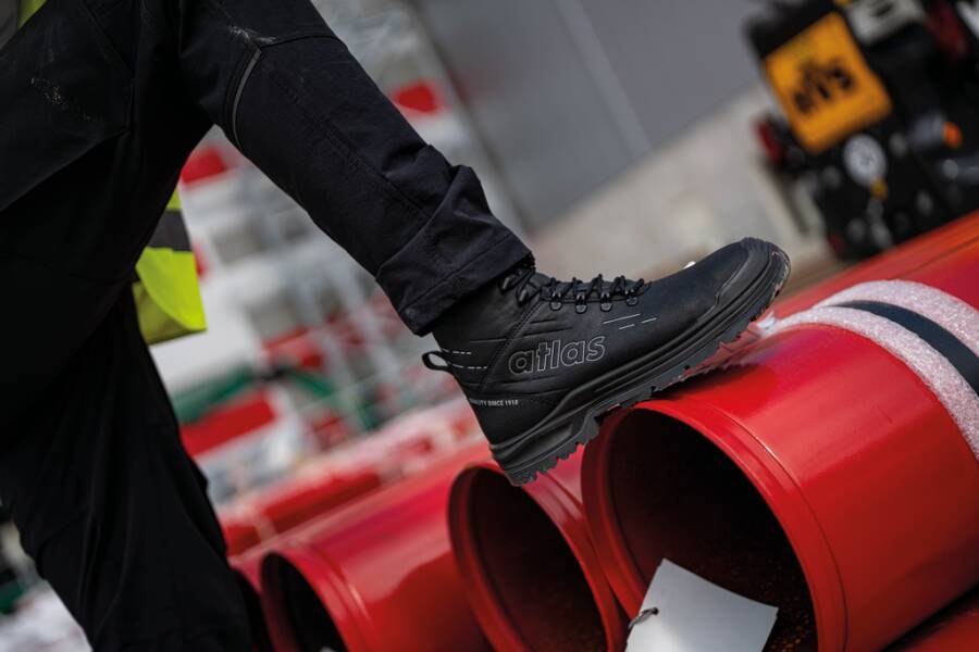 Arbeiter in schwarzen Hosen und Stiefeln tritt auf ein rotes zylindrisches Rohr in einer industriellen Umgebung, um dessen Stabilität während der Handhabung oder Installation zu überprüfen.