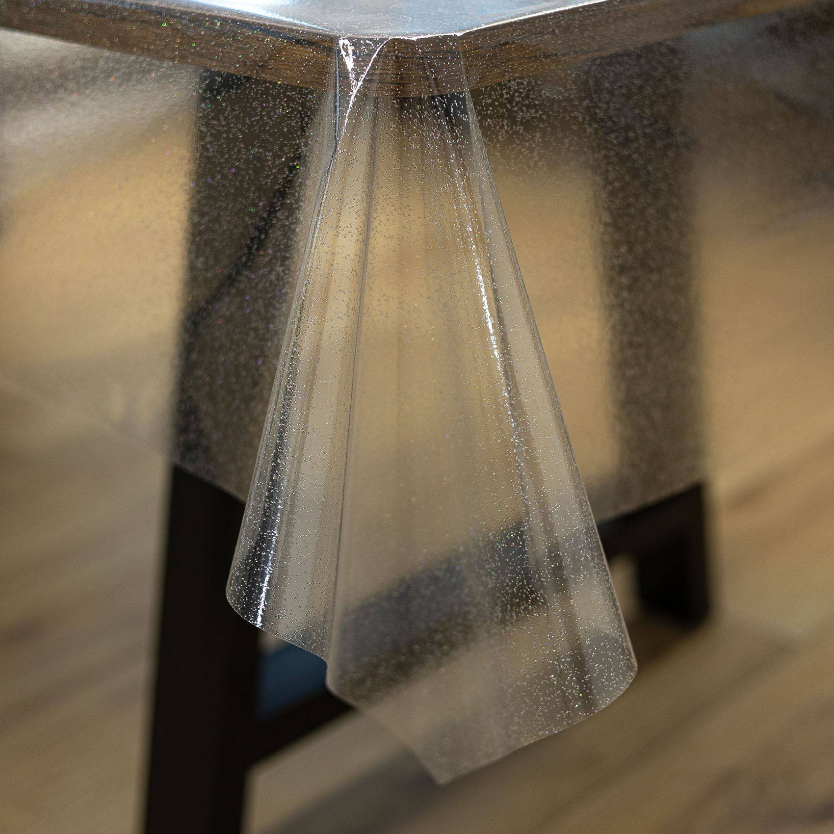 Nahaufnahme einer durchsichtigen Kunststofftischdecke, die von einer Tischkante herunterhängt, mit schwarzen Tischbeinen und einem Holzboden im Hintergrund.