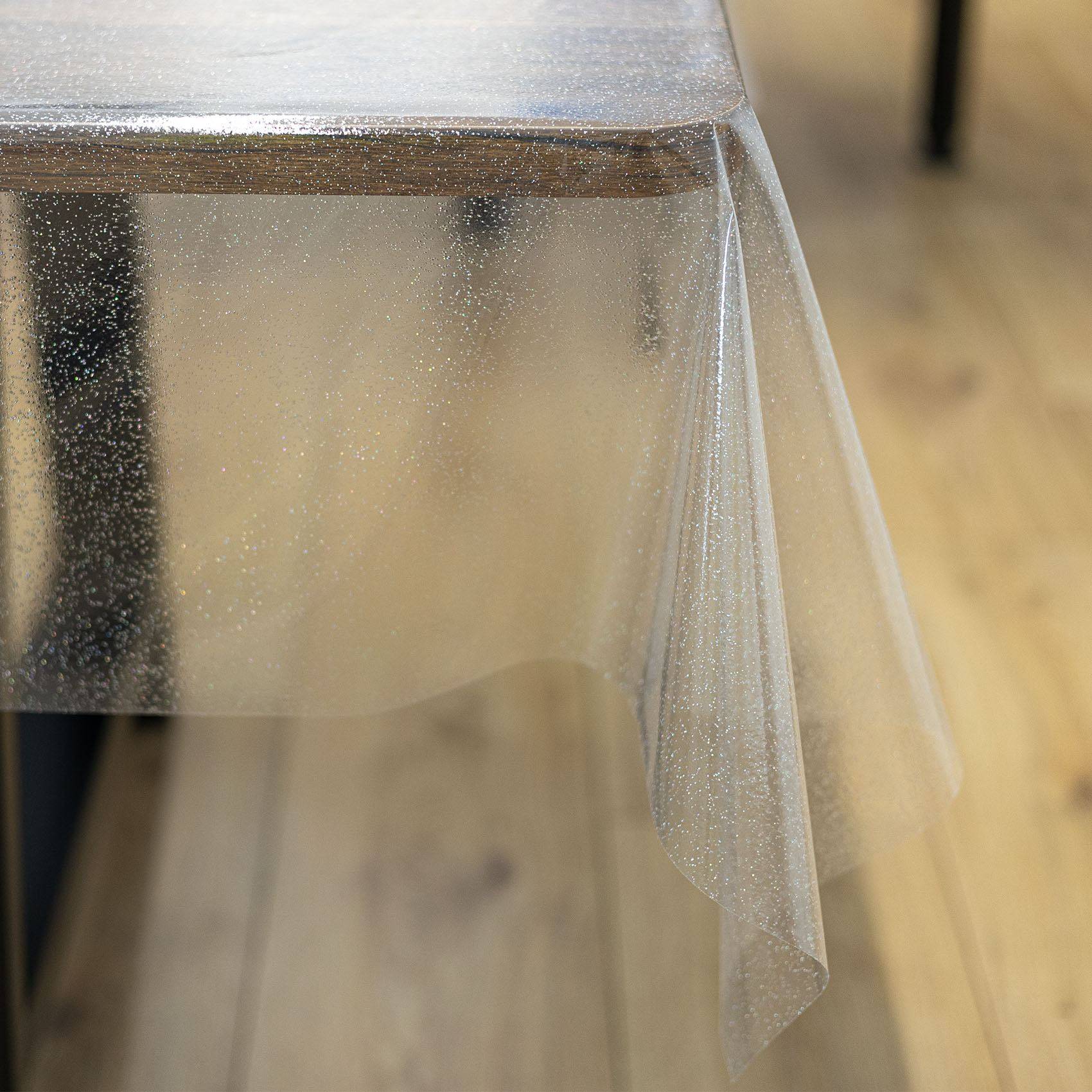 Nahaufnahme einer durchsichtigen Kunststofftischdecke, die über die Ecke eines Tisches drapiert ist, mit dem Holzfußboden im Hintergrund sichtbar.
