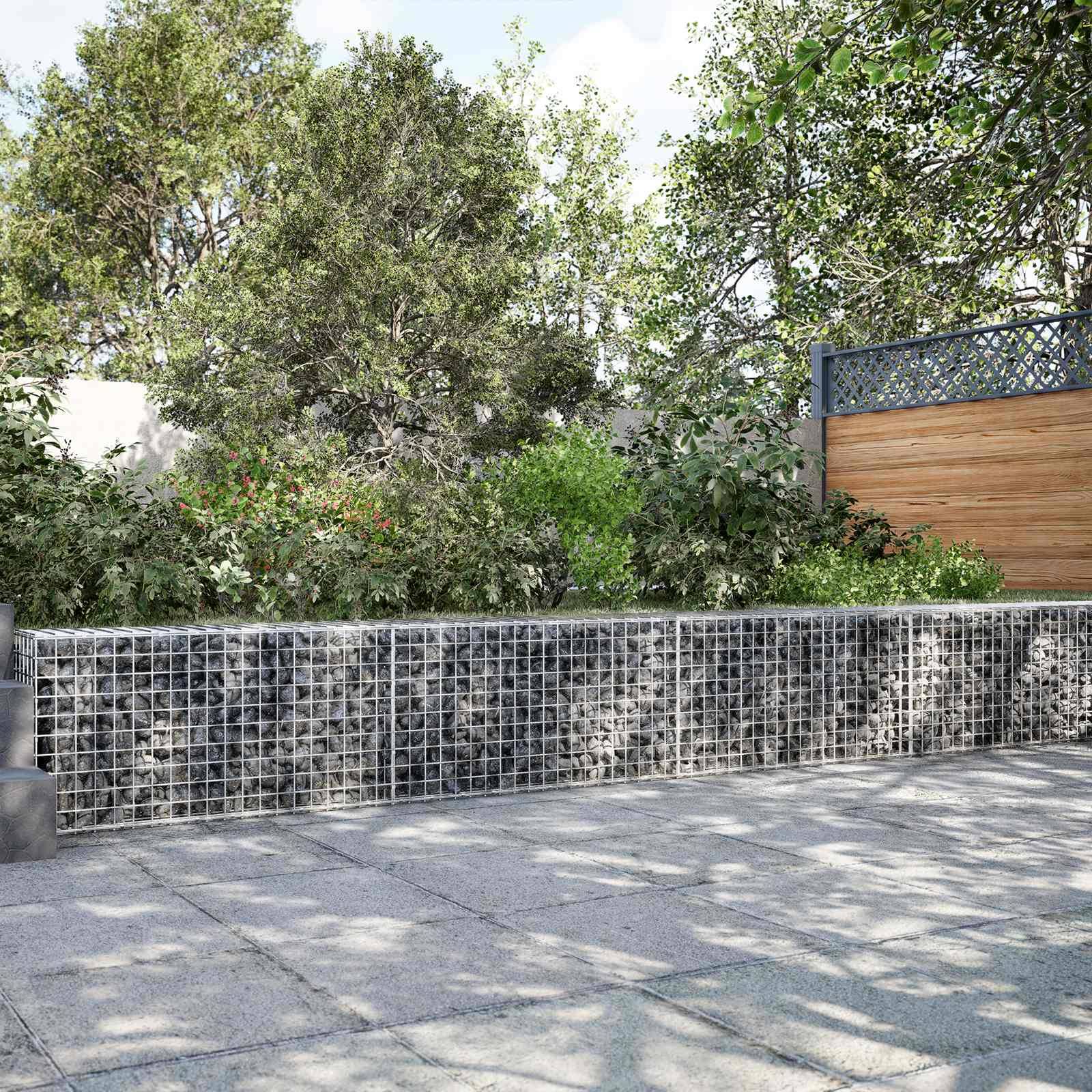 Eine Schotterkorbstützmauer grenzt an einen angelegten Garten mit Bäumen, Sträuchern, einem Holzzaun und einem sonnigen gepflasterten Weg.