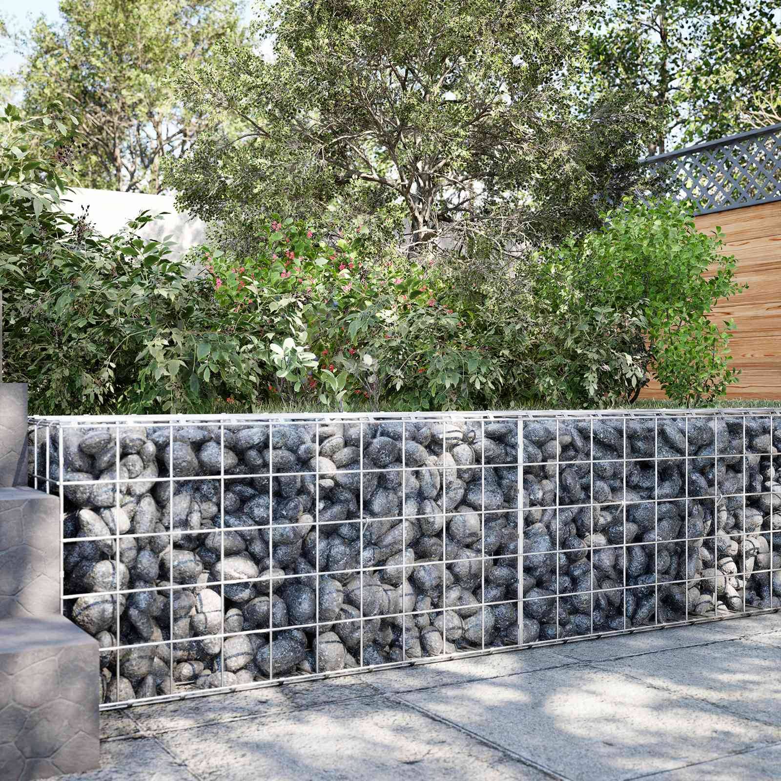 Eine mit Steinen gefüllte Drahtkorbwand grenzt einen Garten mit dichten Bäumen und Sträuchern neben einem gepflasterten Gehweg und einem Holzzaun ab.