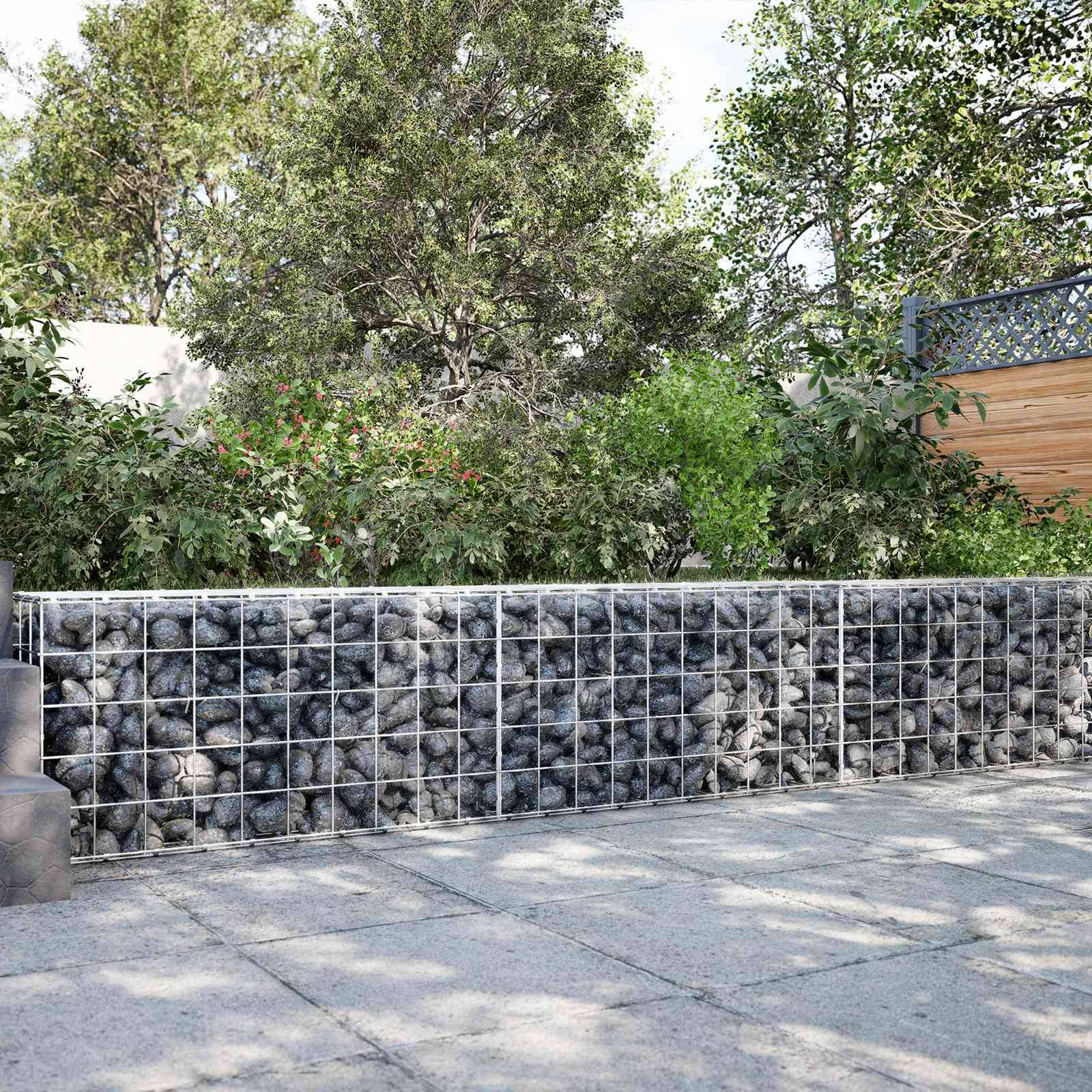 Steingefüllte Drahtgabionenmauer entlang eines befestigten Gehwegs, mit dichtem Baumbestand und Sträuchern dahinter.