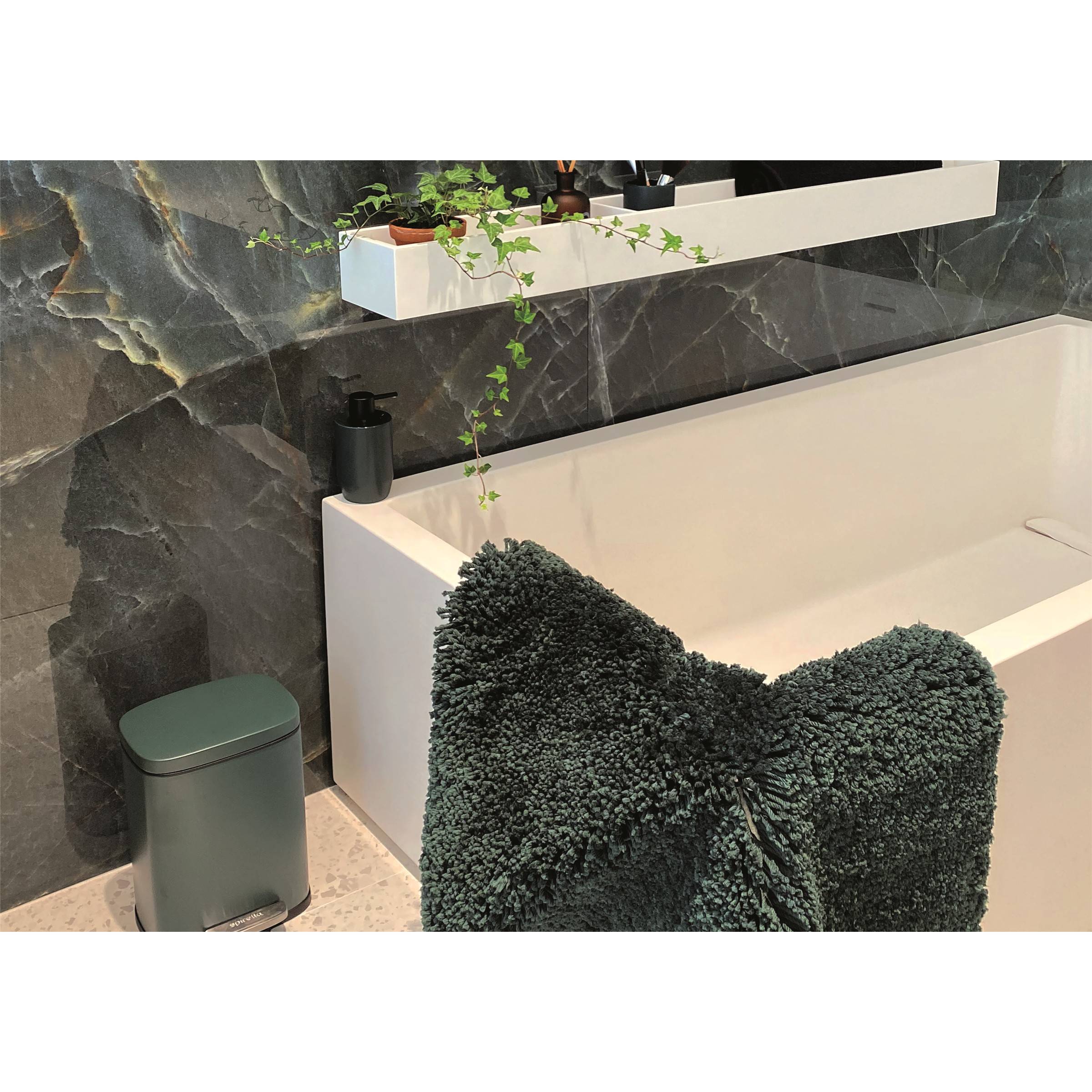 Modernes Badezimmer mit einer weißen Badewanne, dunkler Steinwand, schwebenden Regalen mit Pflanzen und einem dunkelgrünen strukturierten Handtuch oder Stuhl im Vordergrund.