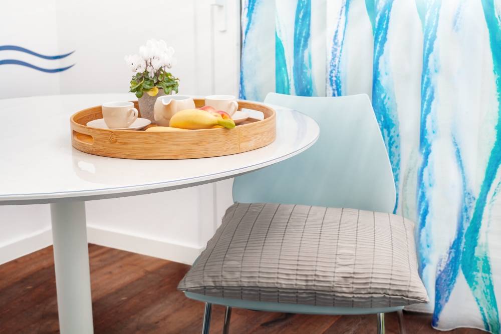 Eine gemütliche Frühstücksecke mit einem weißen runden Tisch, einem Holztablett mit Obst und Tassen sowie einem blaugepolsterten Stuhl neben durchscheinenden blauen Vorhängen.
