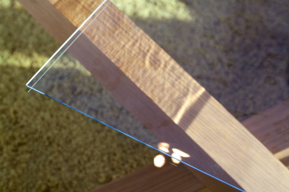 Nahaufnahme einer Glastischplatte-Ecke mit einem Holzbalken, der sich darin spiegelt, beleuchtet von Sonnenlicht und mit weichen Schatten.