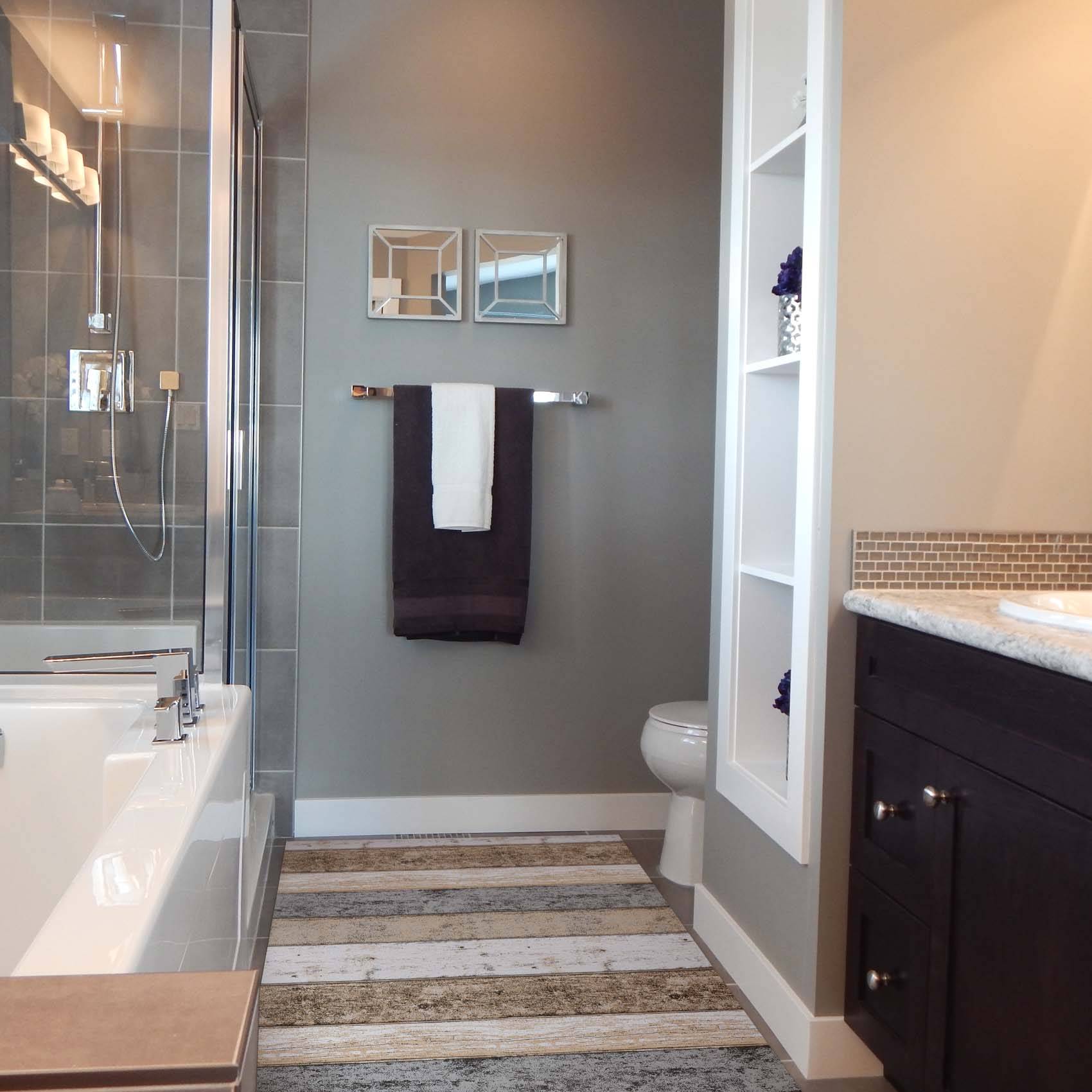 Modernes Badezimmer mit Glasdusche, Badewanne, Waschtisch, Toilette und gestreiftem Holzlook-Boden; graue Wände mit Handtuch und gerahmten Spiegeln.