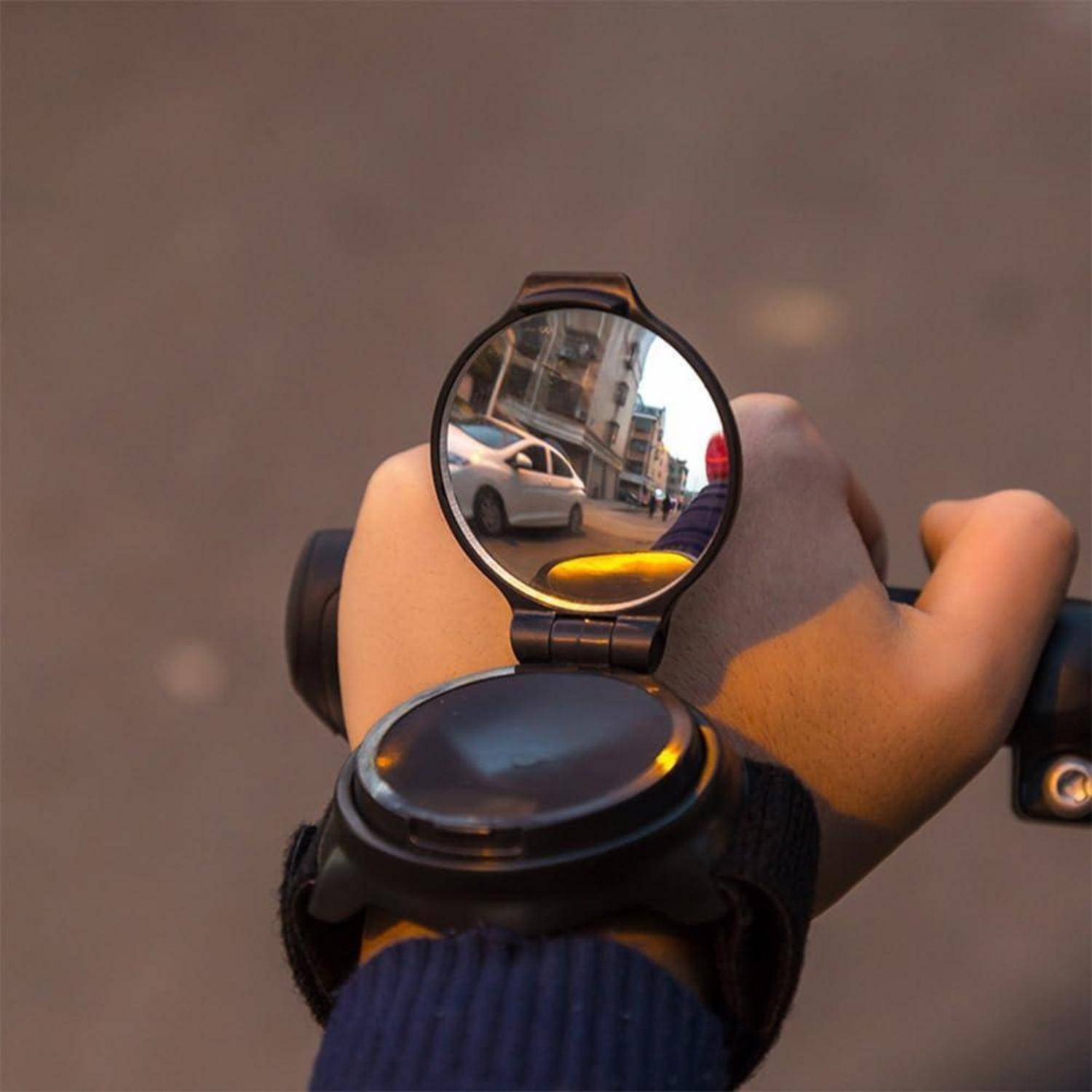 Nahaufnahme der Hand eines Radfahrers am Lenker mit einem runden Rückspiegel, der eine Straße und Autos dahinter reflektiert.