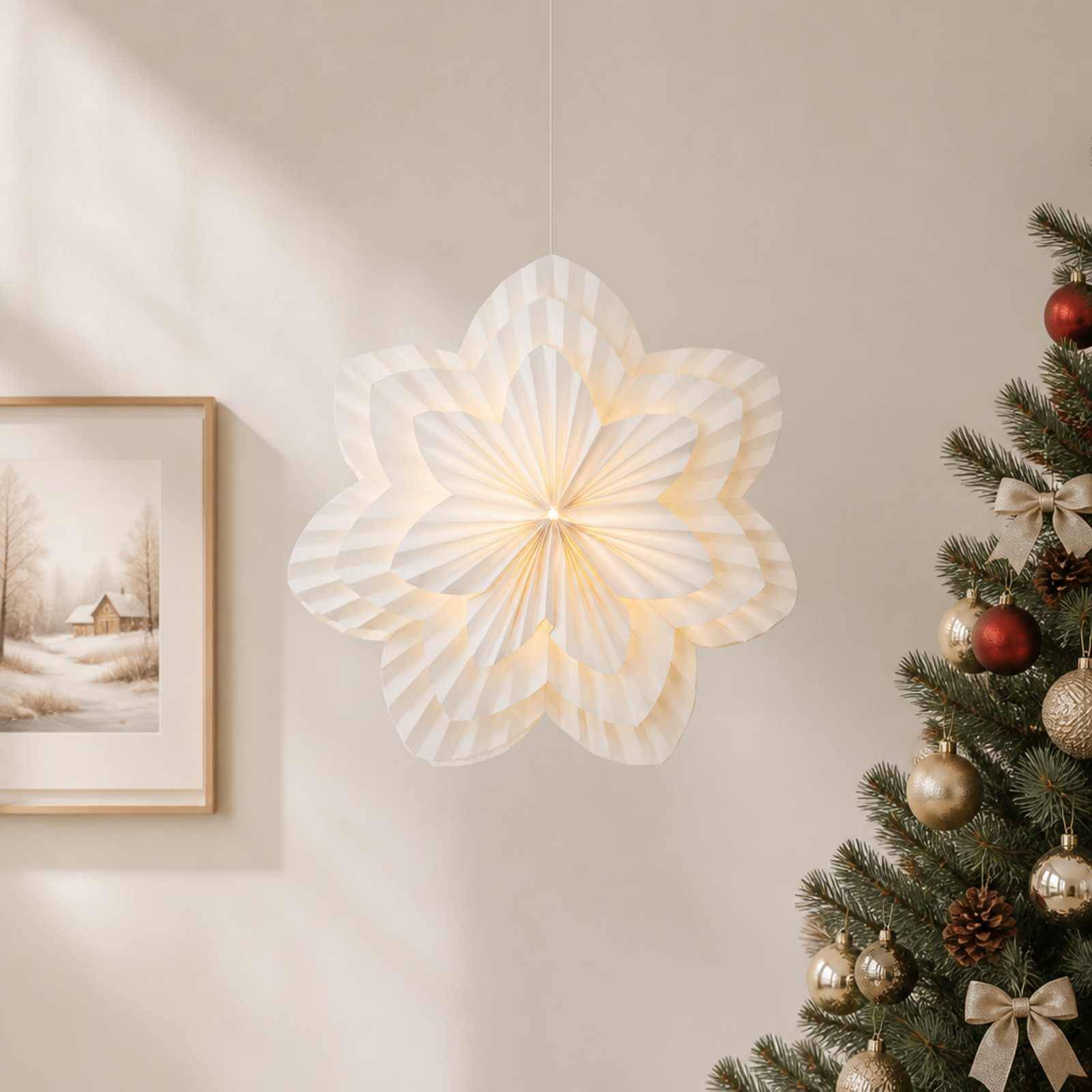 Eine leuchtende weiße Papiersternlaterne hängt an einer Wand neben einem gerahmten Winterdruck und einem geschmückten Weihnachtsbaum.