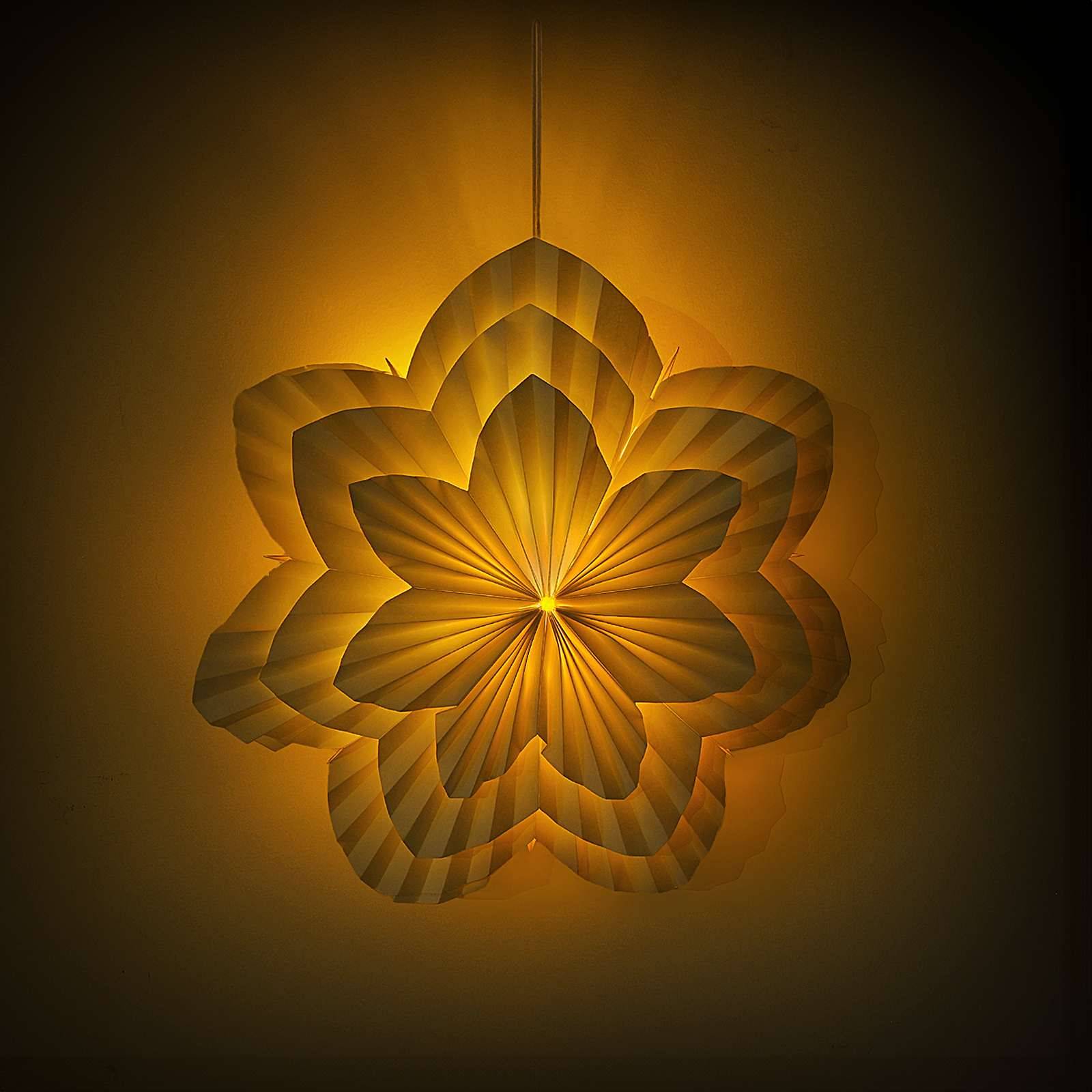 Hängende dekorative Papierllampe in Form einer geschichteten Blume, die gegen einen dunklen Hintergrund in warmem Bernsteinton leuchtet.