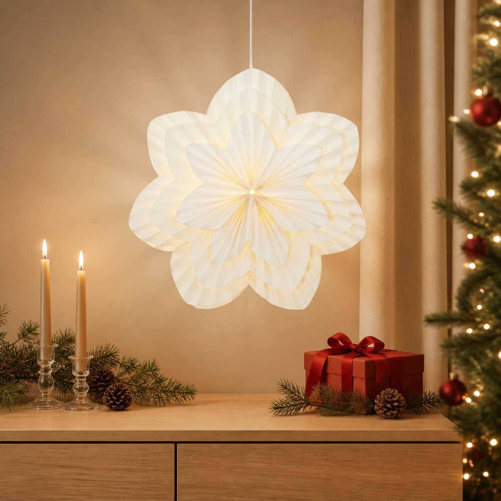 Warme Weihnachtsszene mit einer leuchtenden Papierschneeflocken-Laterne, zwei brennenden Kerzen, Tannenzweigen und Zapfen auf einem Holzschrank, und einer roten Geschenkbox neben einem geschmückten Baum.