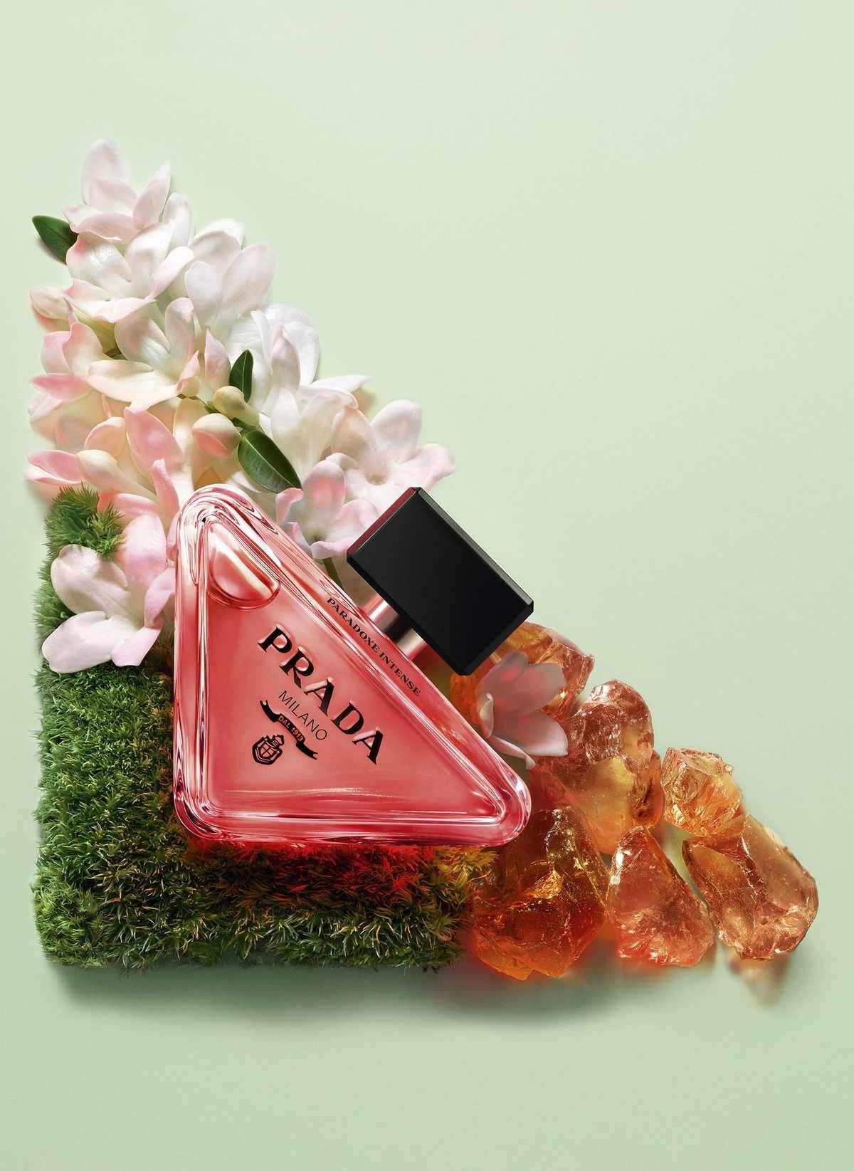 Prada Paradoxe Parfümflasche auf Moos mit weißen Blüten und Bernsteinkristallen vor hellgrünem Hintergrund.
