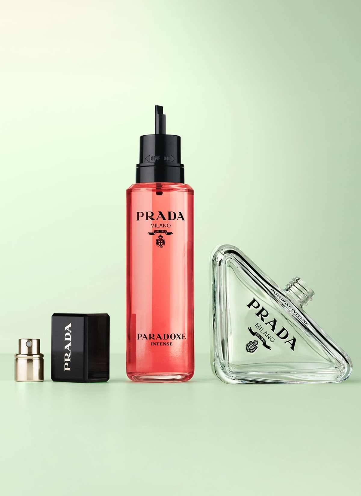 Drei Prada-Duftflaschen und eine Kappe auf einem blassgrünen Hintergrund präsentiert, einschließlich einer hohen roten Sprühflasche und einer dreieckigen klaren Flasche.