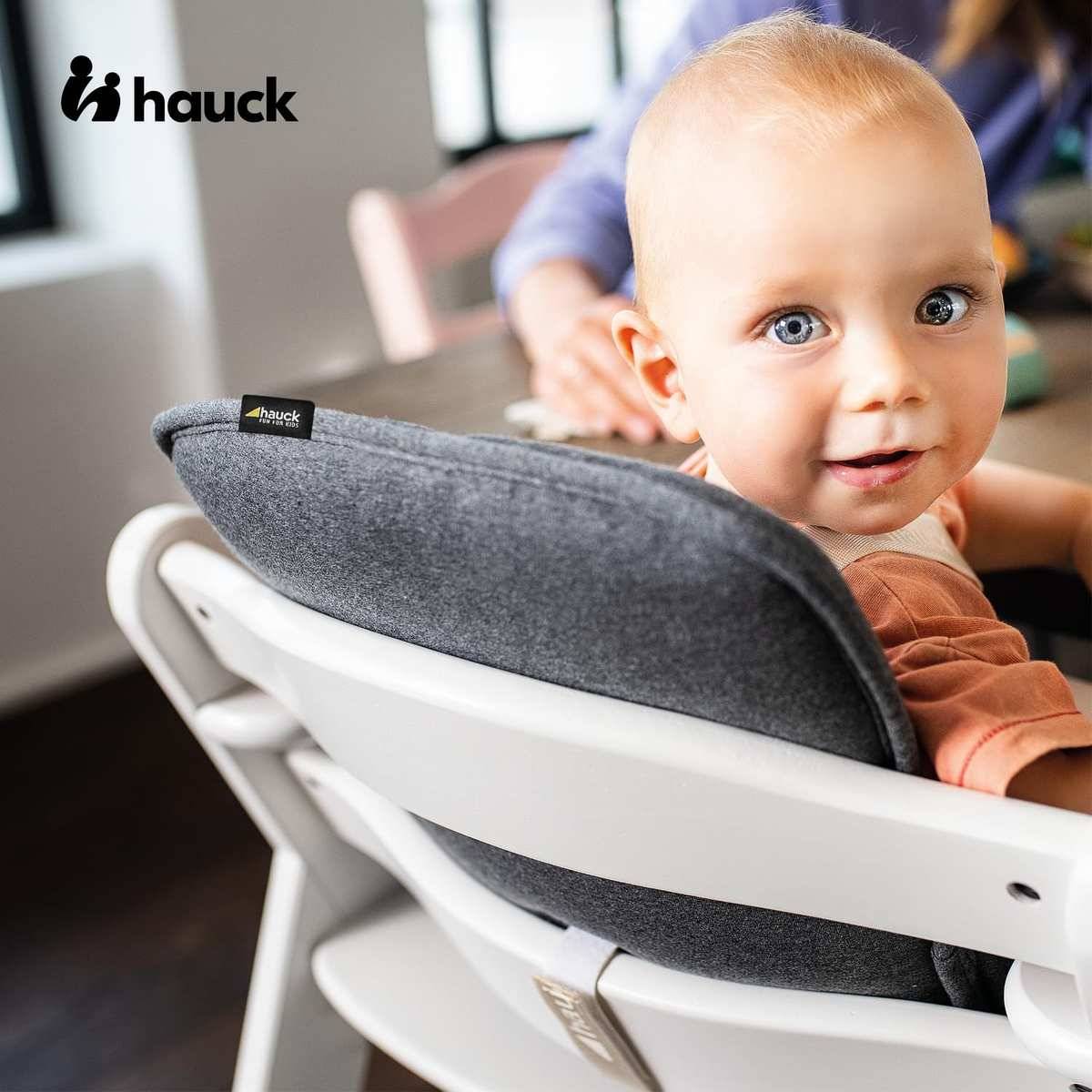 Lächelndes Baby sitzt in einem weißen Hochstuhl mit grauem Kissen und schaut in die Kamera; das Logo "hauck"" erscheint oben links.