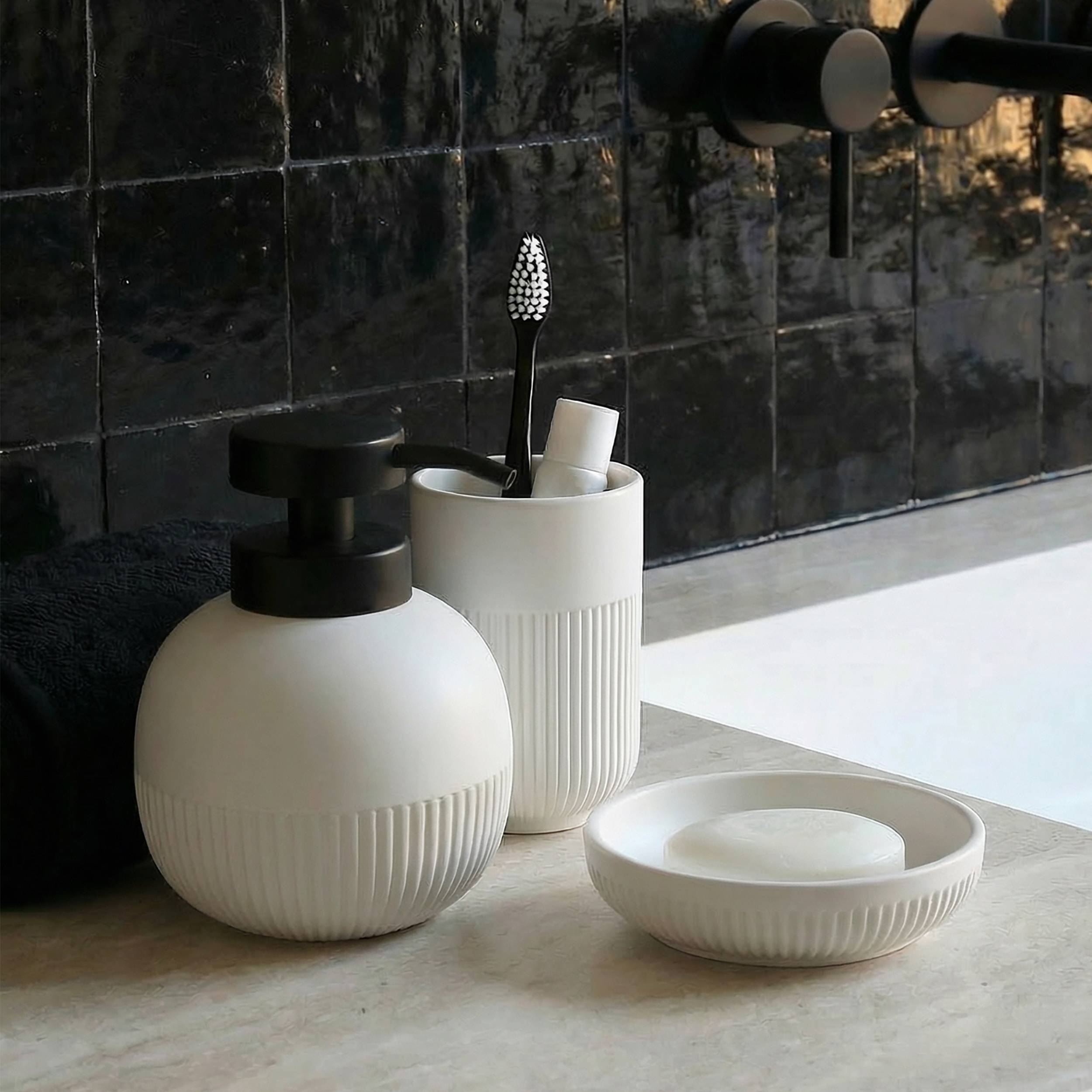 Moderne Badezimmeraccessoires auf einer Arbeitsplatte: ein Seifenspender, ein Zahnbürstenhalter und eine Seifenschale vor einer dunklen gefliesten Wand.