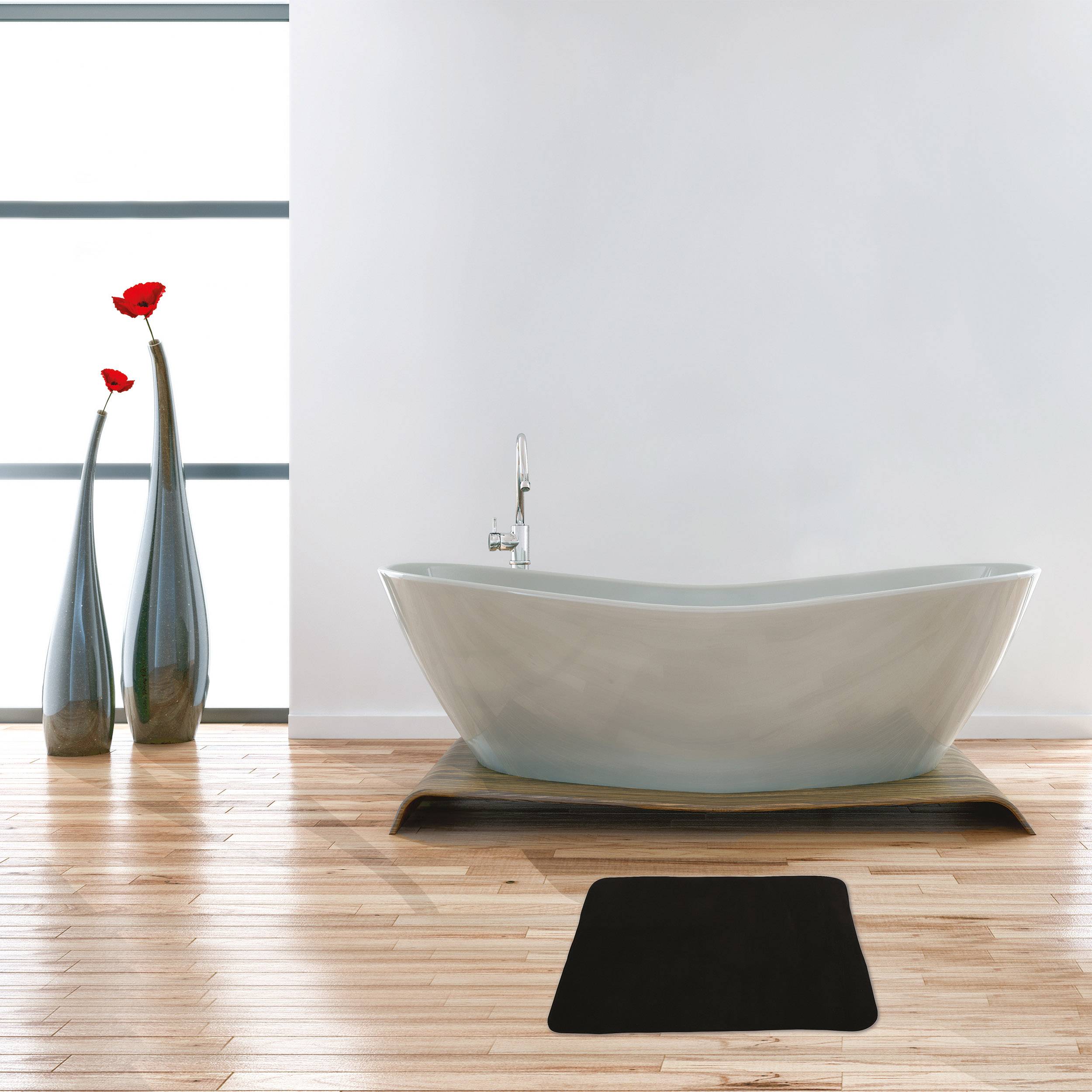 Modernes Badezimmer mit einer freistehenden weißen Badewanne in der Mitte auf einem Holzboden, schwarze Badematte davor und zwei hohe Vasen mit roten Blumen am Fenster.