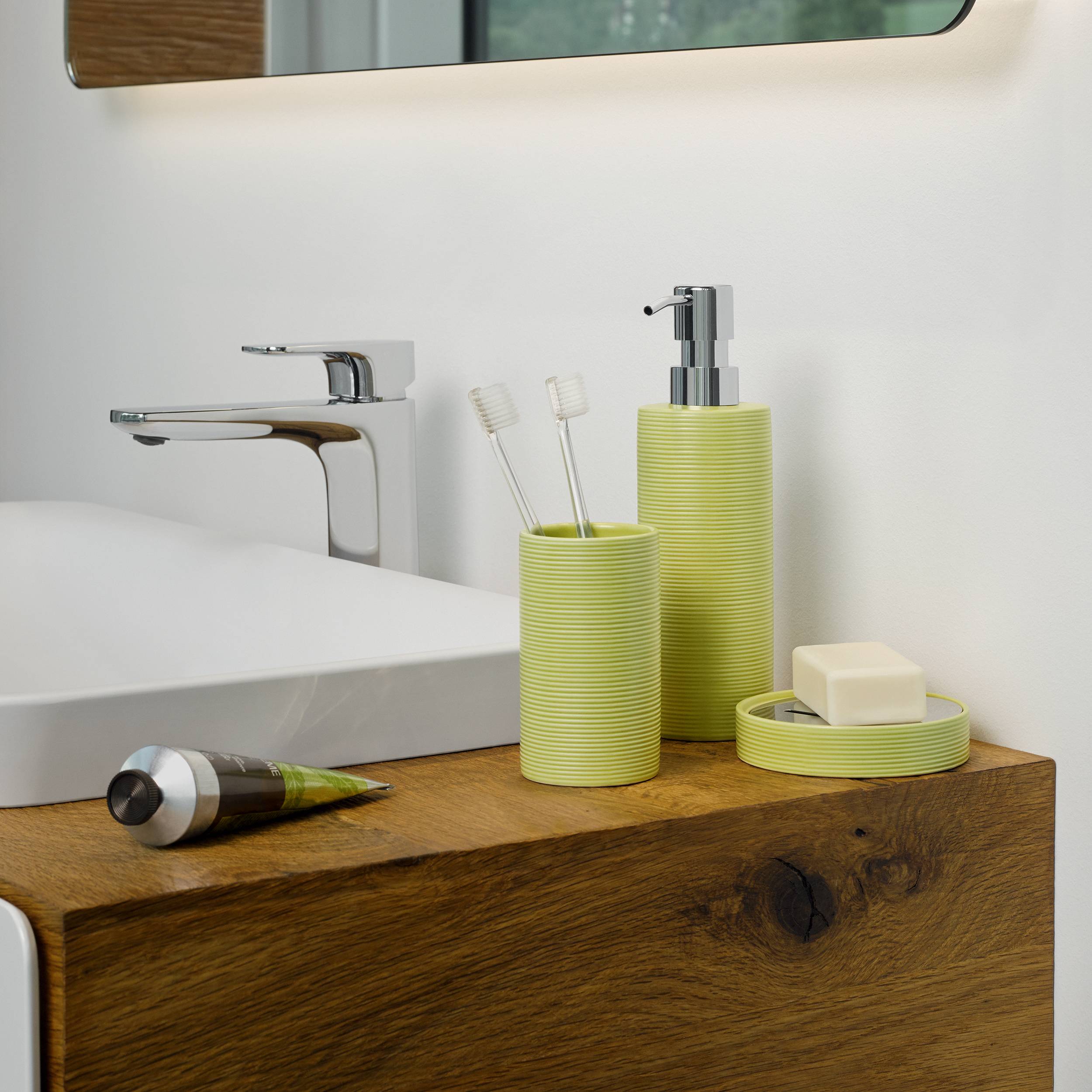 Modernes Badezimmerwaschtisch mit Waschbecken und Chromwasserhahn, limegrüner Seifenspender und Zahnbürstenhalter, Seifenschale mit Seifenstück und Zahnpastatube auf Holzablage.