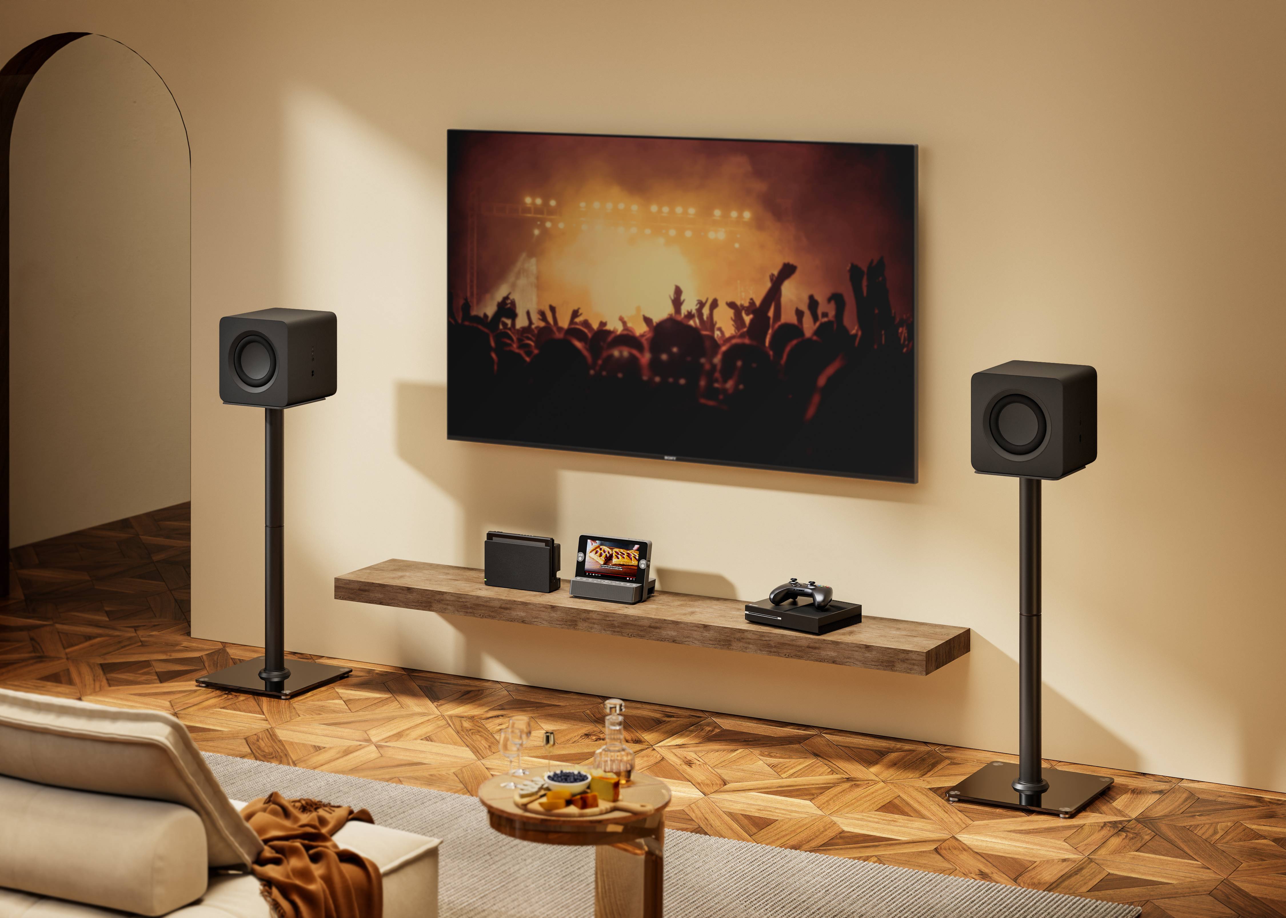 Modernes Wohnzimmer mit an der Wand montiertem Fernseher, der eine Konzertmenge zeigt, flankiert von zwei Lautsprechern auf Ständern, mit einem schwebenden Regal darunter, das Mediengeräte enthält.