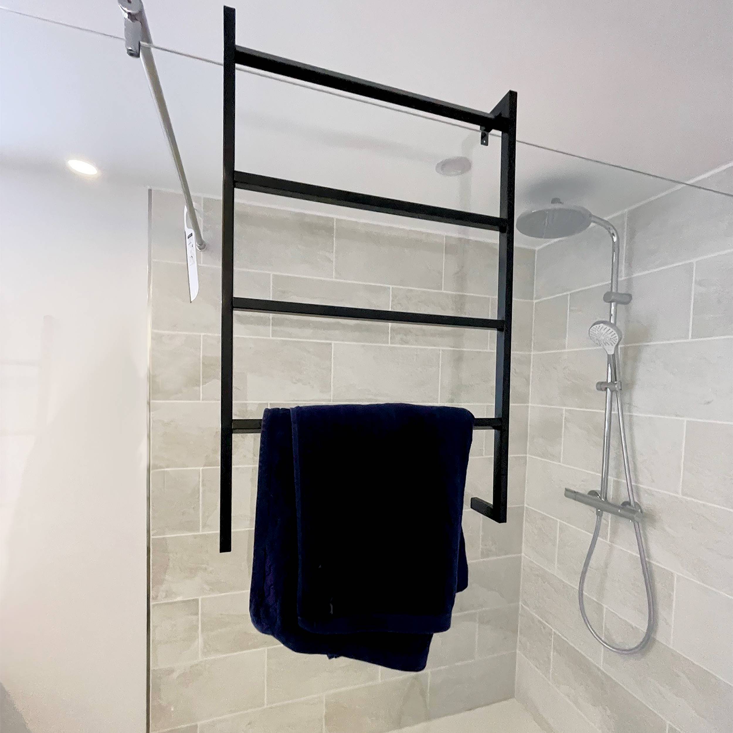 Modernes gekacheltes Badezimmer mit einem schwarzen Handtuchhalter im Leiterstil, der über die Glasumrandung hängt und ein dunkelblauesi Handtuch hält, neben einem Chrombrausekopf und einem Schlauch.