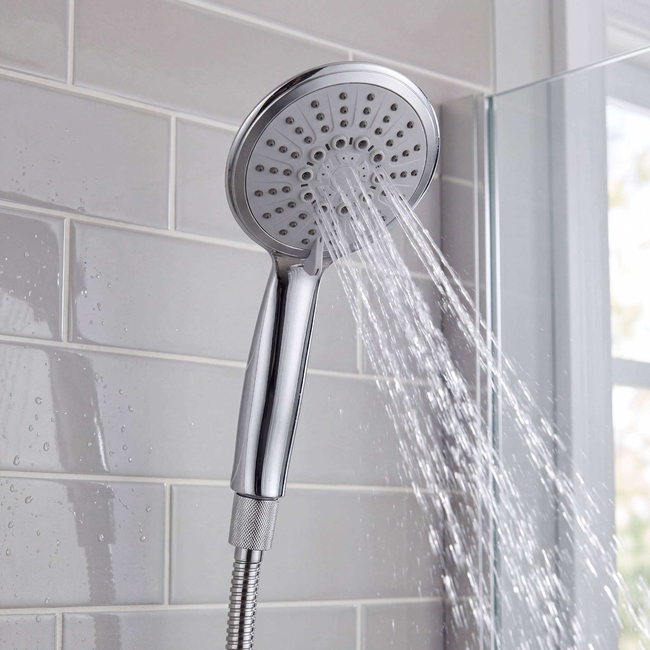Verchromter tragbarer Duschkopf sprüht Wasser in einer gefliesten Duschkabine neben einer Glastür.