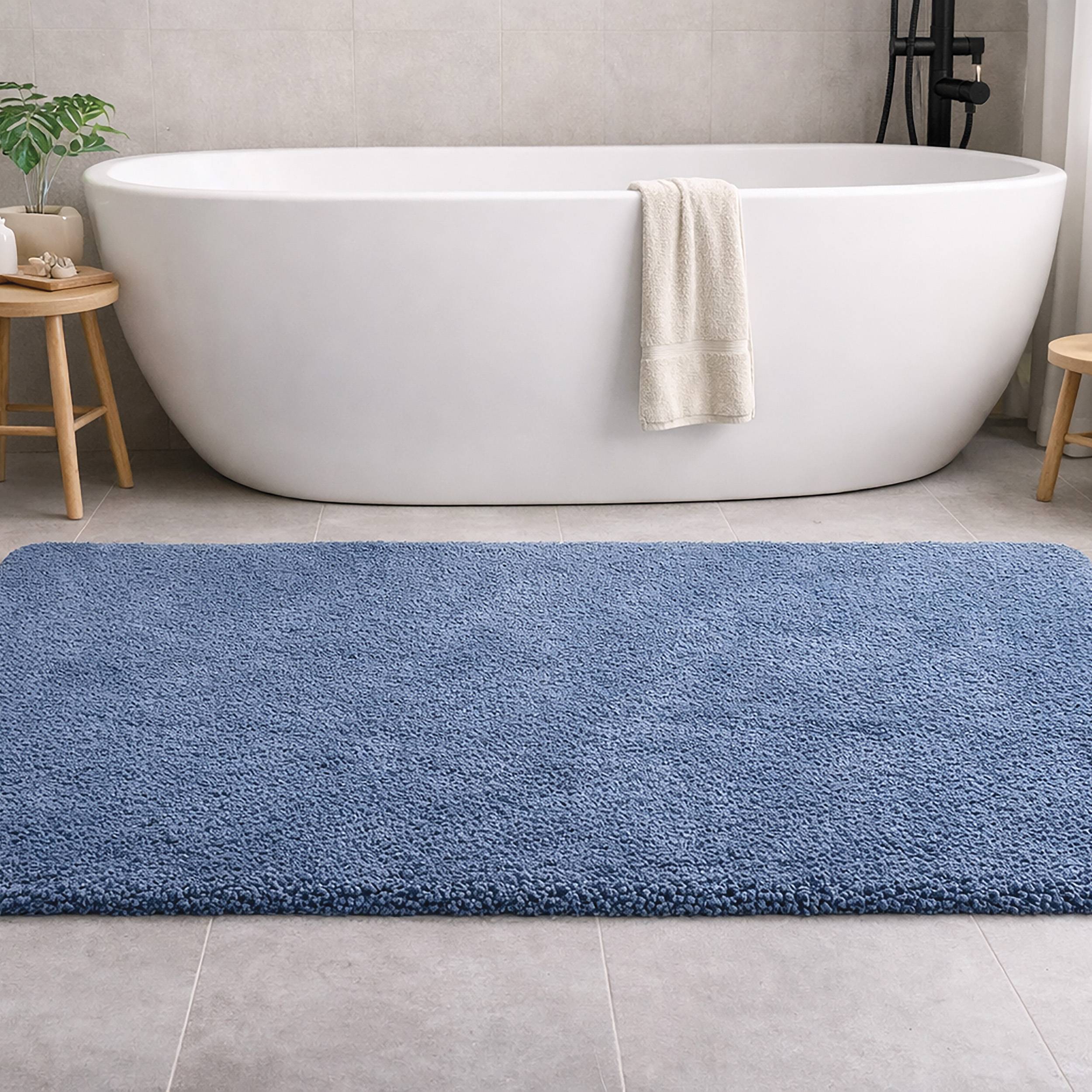 Modernes Badezimmer mit einer freistehenden weißen Badewanne, ein über der Vorderseite drapiertes Handtuch, flankiert von kleinen Holzhockern und einem großen blauen Badteppich.