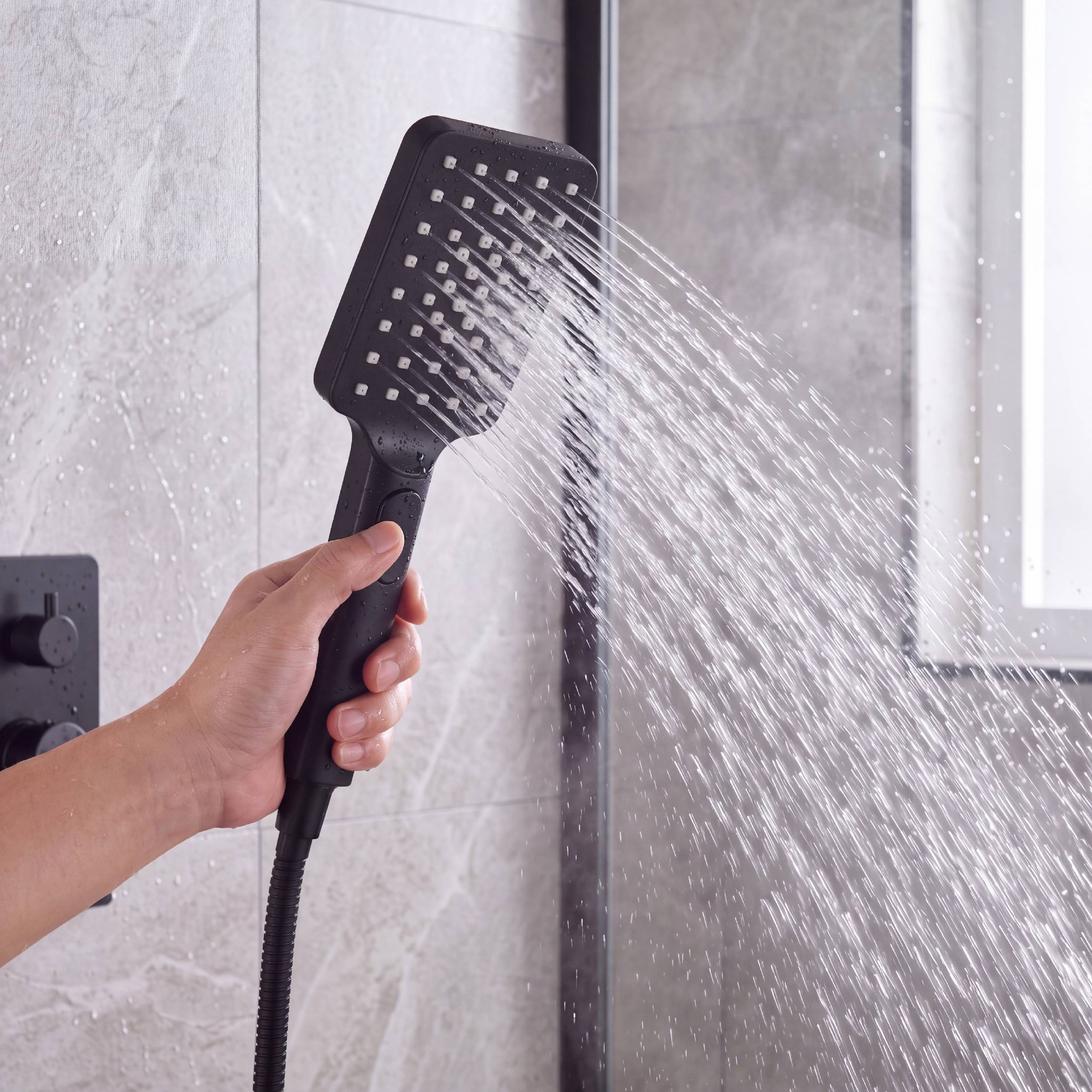 Hand hält einen schwarzen tragbaren Duschkopf und sprüht Wasser in einer Duschkabine im Badezimmer.