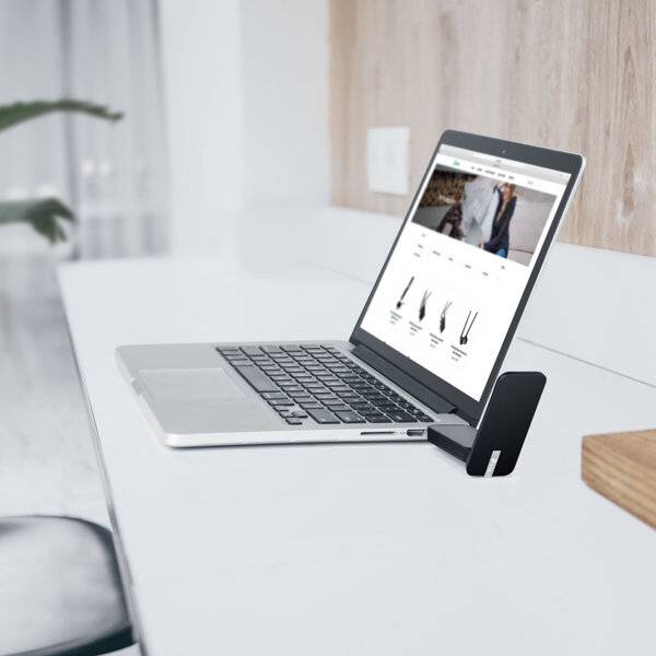 Laptop auf einem Schreibtisch geöffnet, das eine Online-Shopping-Website mit Kleidung und Produktlisten anzeigt, auf einem Telefonständer in einem minimalistischen Büro angesehen.