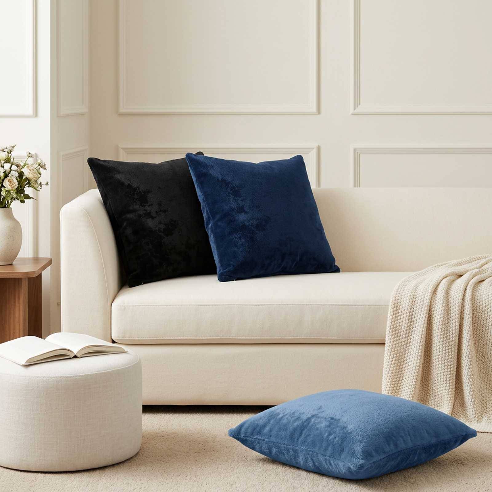 Cremefarbenes Sofa mit schwarzen und marineblauen Kissen in einem hellen Wohnzimmer; blaues Bodenkissen, Hocker, Beistelltisch und drapierte Decke schaffen ein gemütliches, neutrales Interieur.