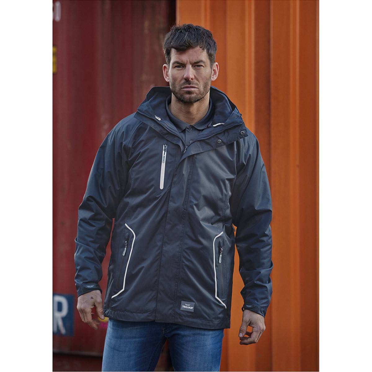 Ein Mann in einer dunklen wasserdichten Jacke und Jeans steht im Freien vor roten und orangefarbenen Schiffscontainern und blickt in die Kamera.