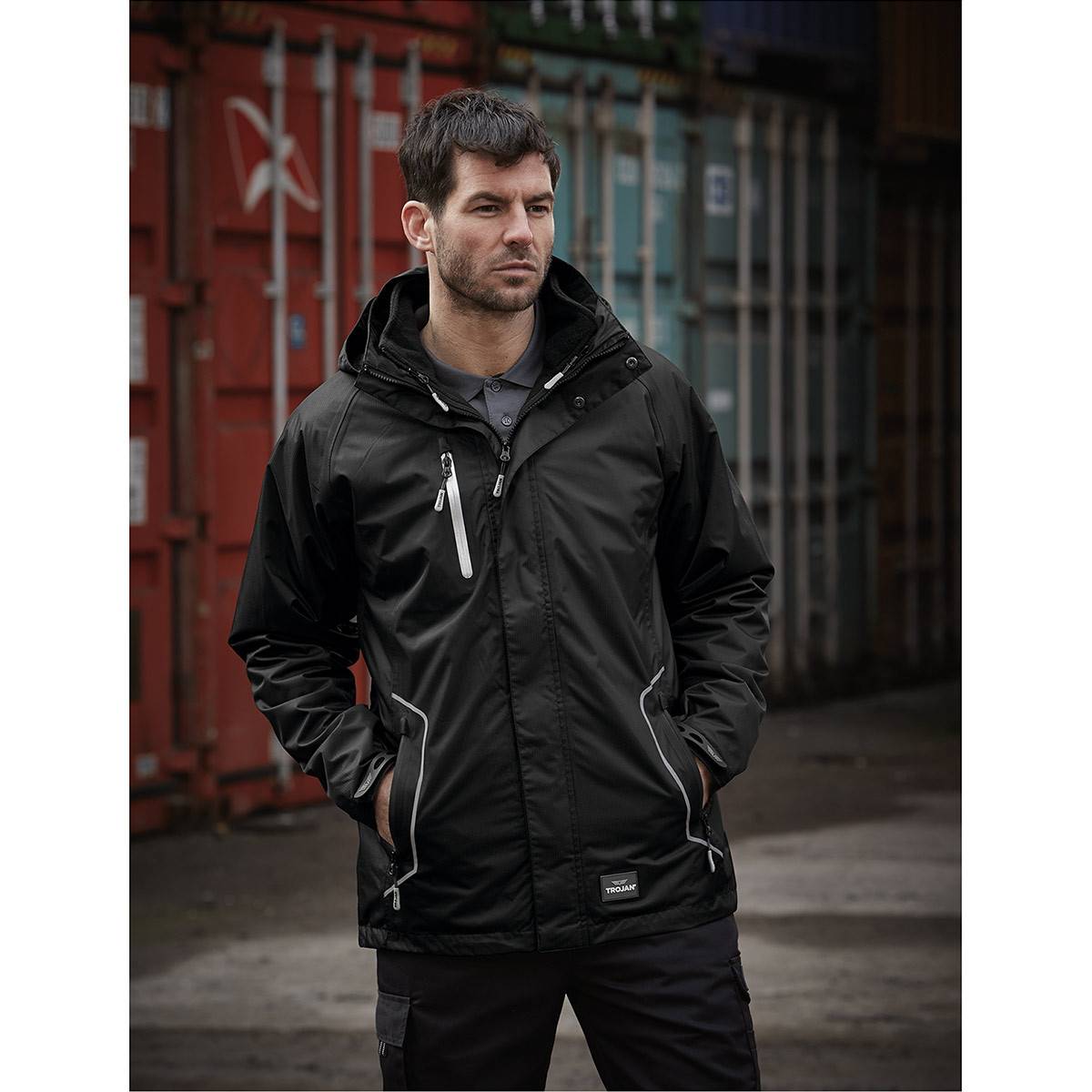 Ein Mann in einer schwarzen Kapuzenjacke steht mit den Händen in den Taschen in einer urbanen Industrieumgebung.