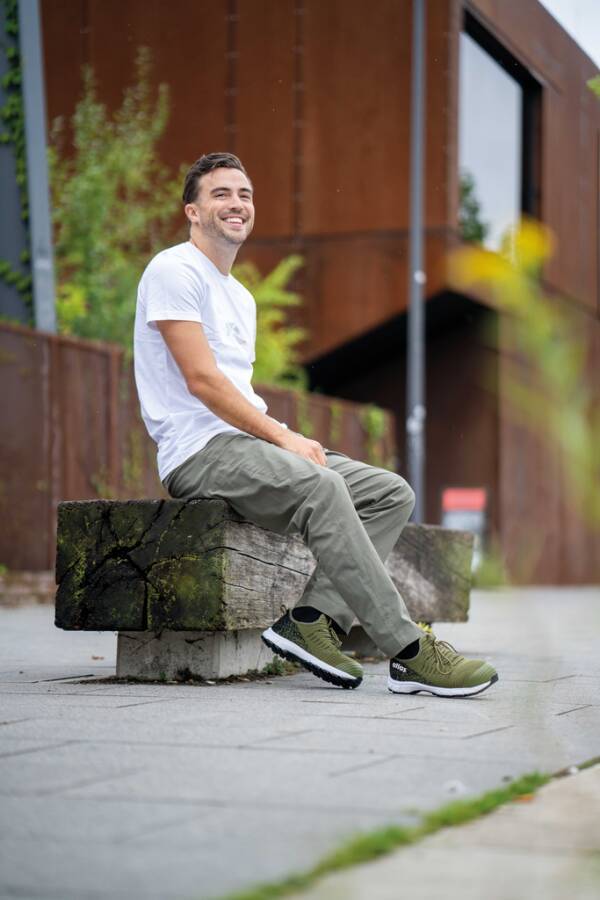 Ein lächelnder Mann in einem weißen T-Shirt und grüner Hose sitzt auf einer Holzbank im Freien in einer urbanen Umgebung.