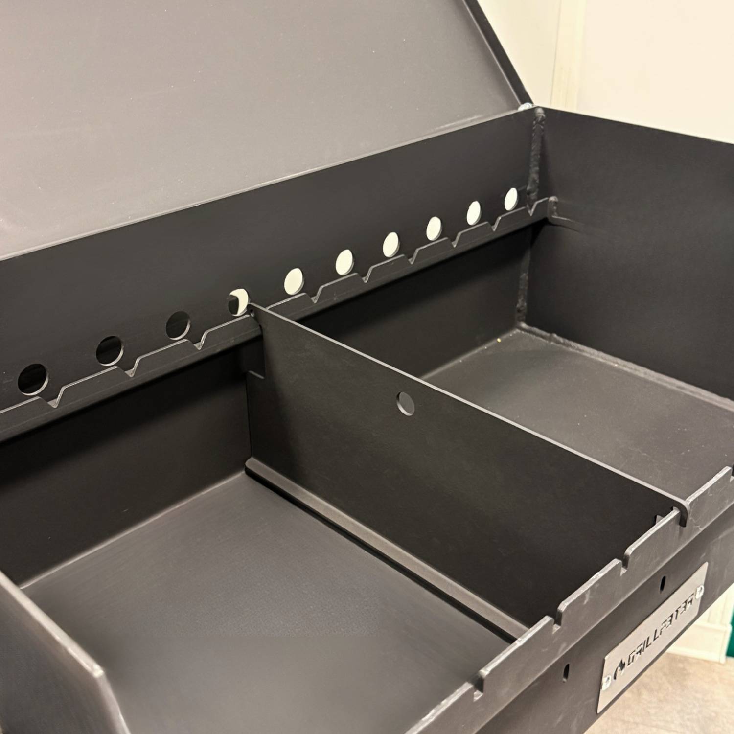 Offene schwarze Aufbewahrungsbox mit zwei geteilten Fächern und seitlichen Ausschnittlaschen, von oben in perspektivischer Ansicht dargestellt.