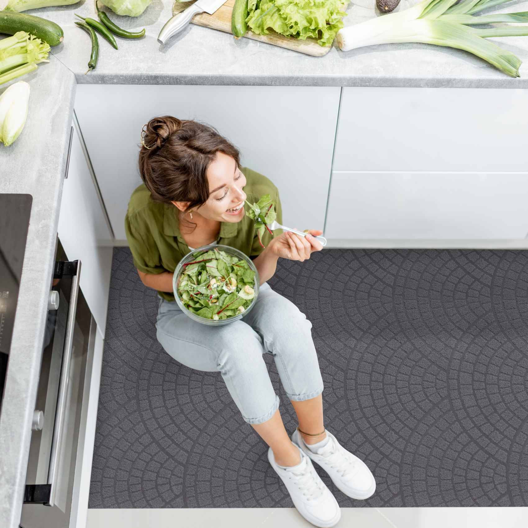 Frau sitzt auf dem Küchenboden und isst einen Salat aus einer Schüssel neben der Arbeitsplatte.