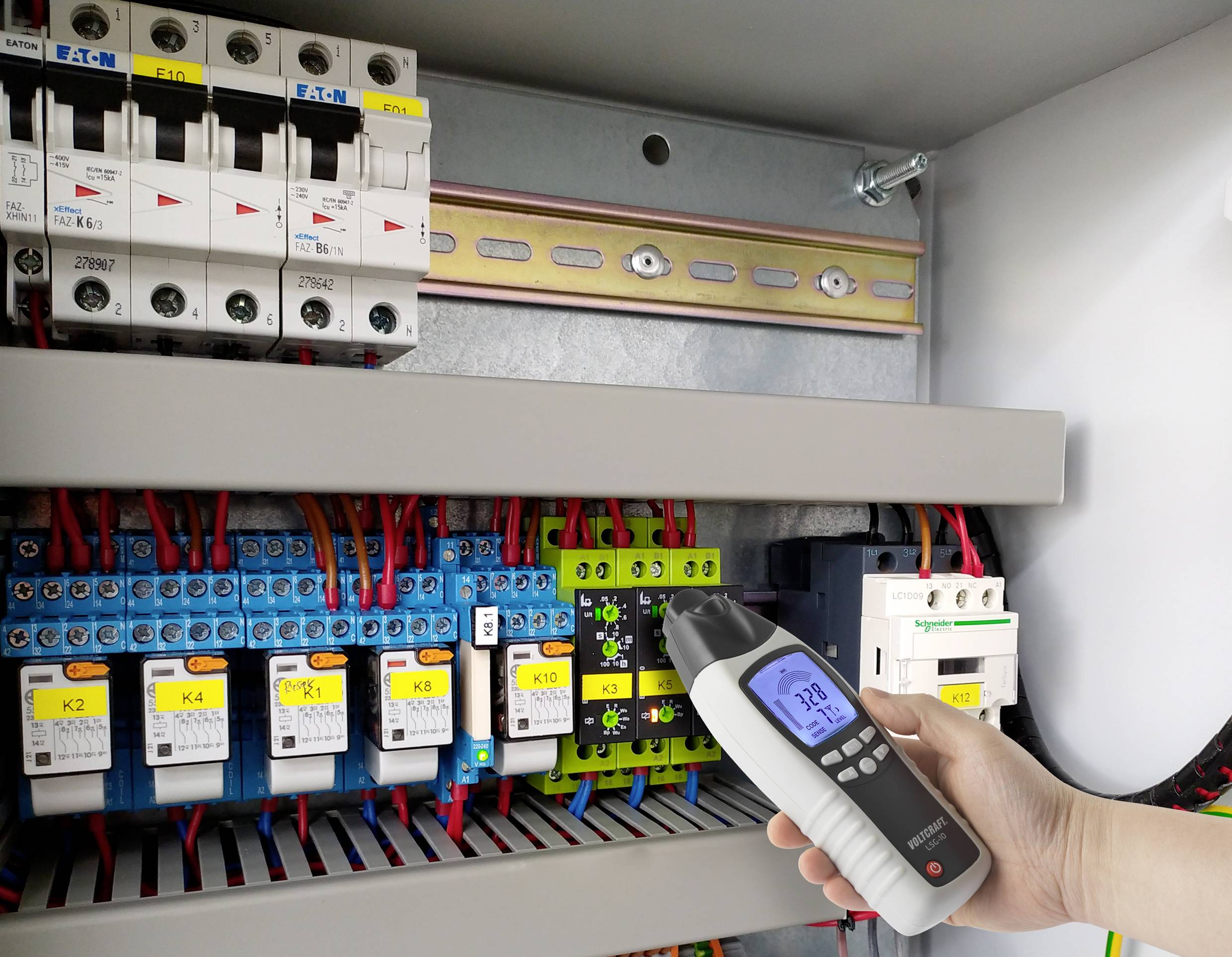 A hand holds a multimeter measuring the voltage in an open electrical cabinet. Various switches and wiring are visible.