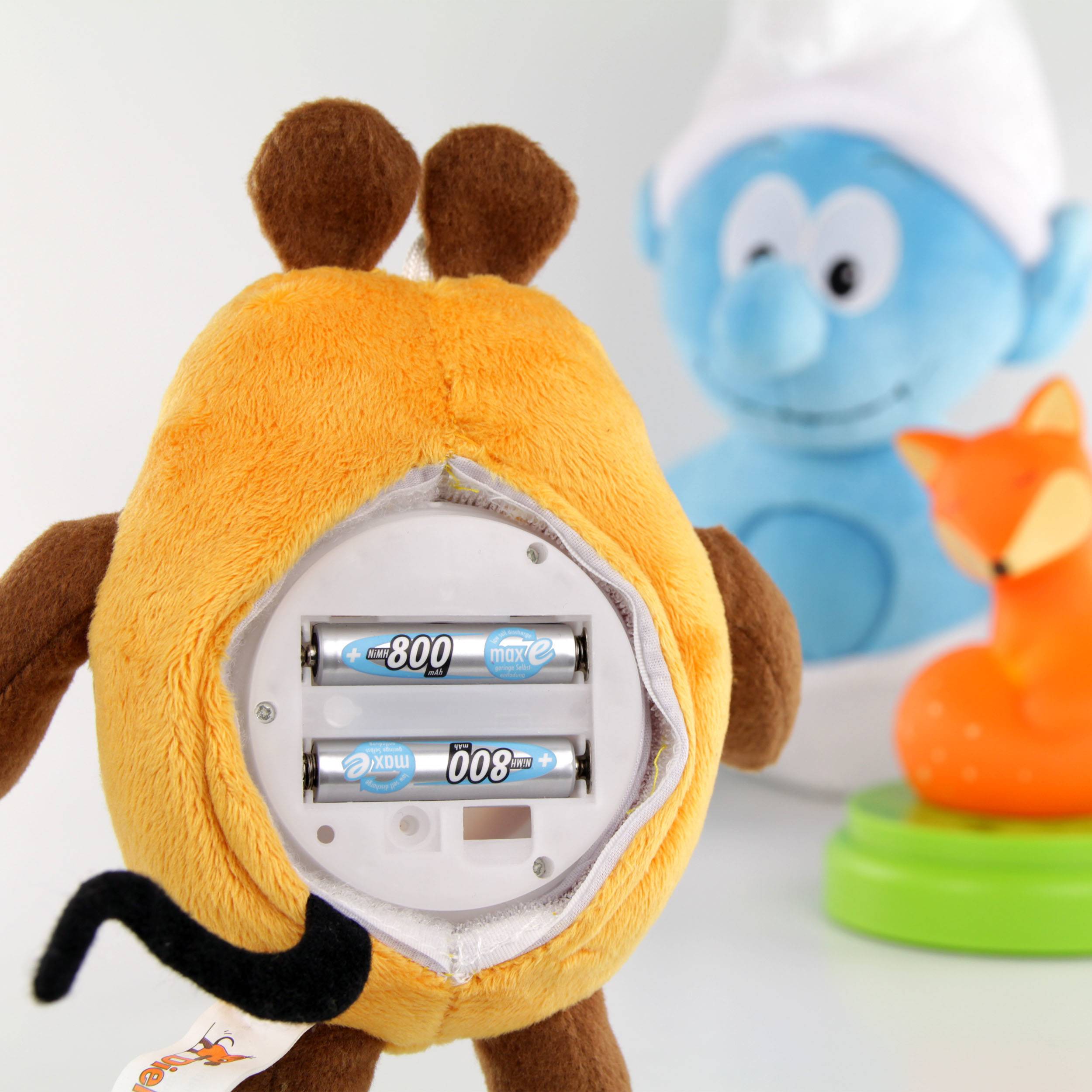 An orange plush toy with removable batteries; behind it a blue doll and an orange toy on a green base.