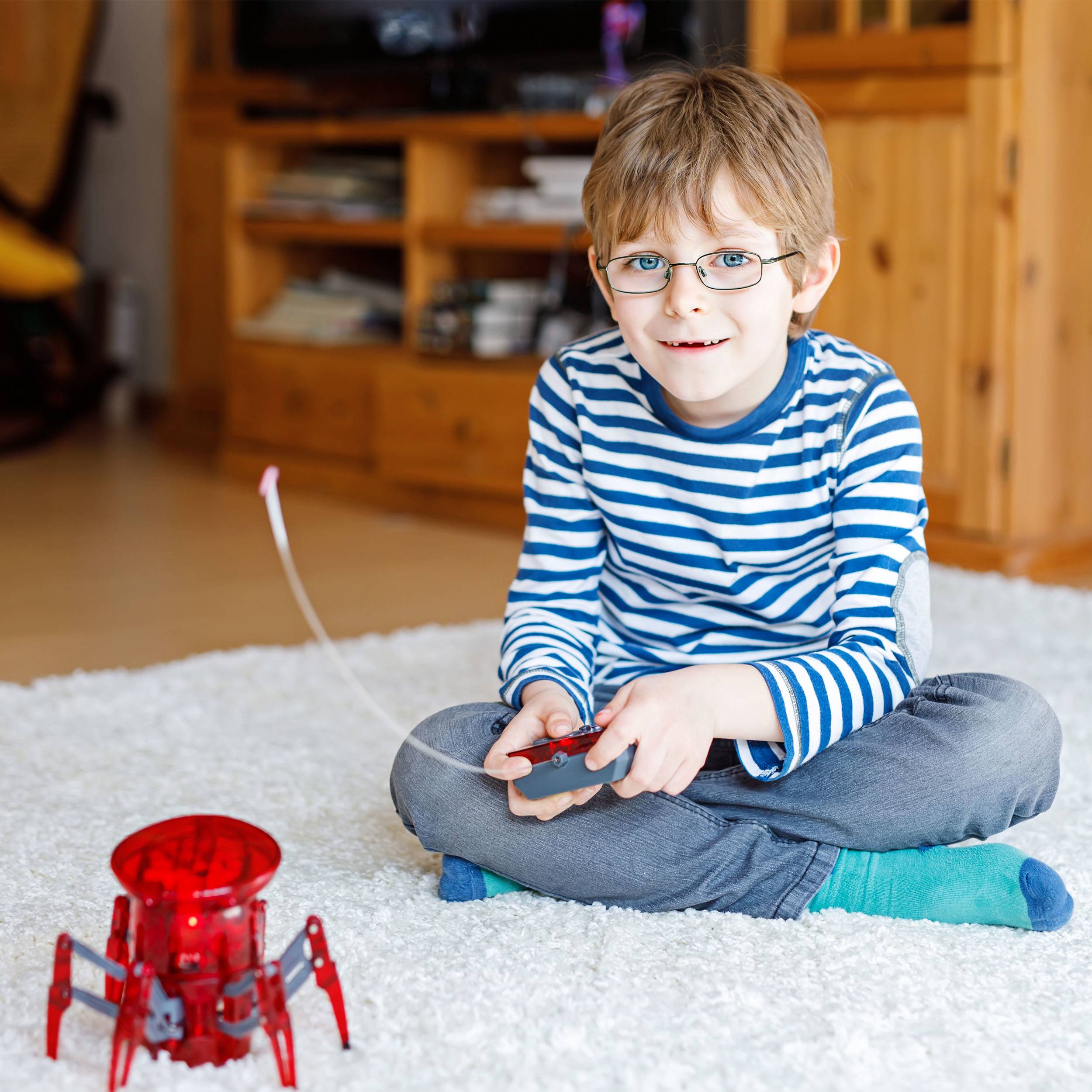 A boy is playing on a carpet with a remote-controlled red robot toy in a living room.