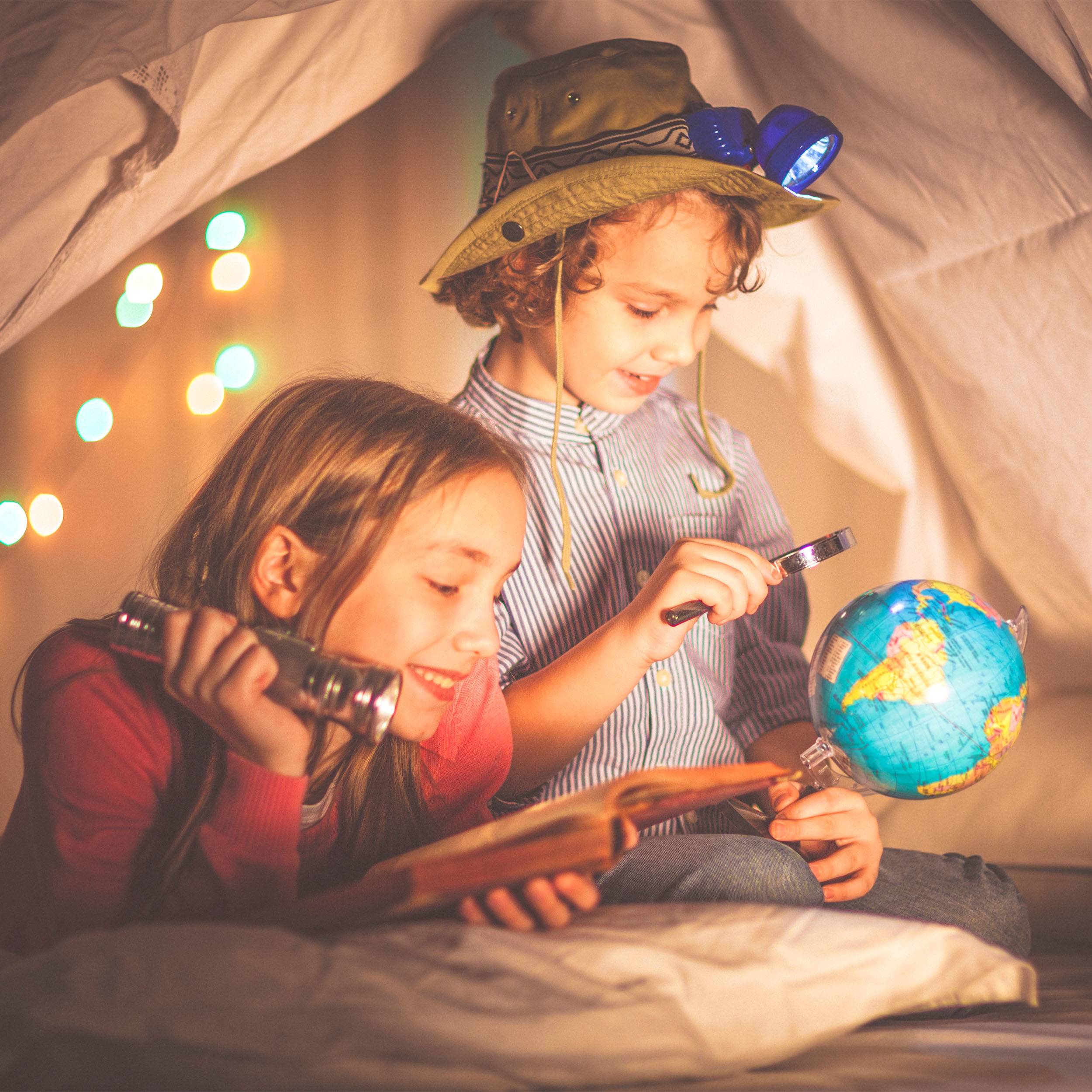 Two children are lying in a tent, curiously examining a book. One is holding a pair of binoculars, while the other is pointing at a globe.